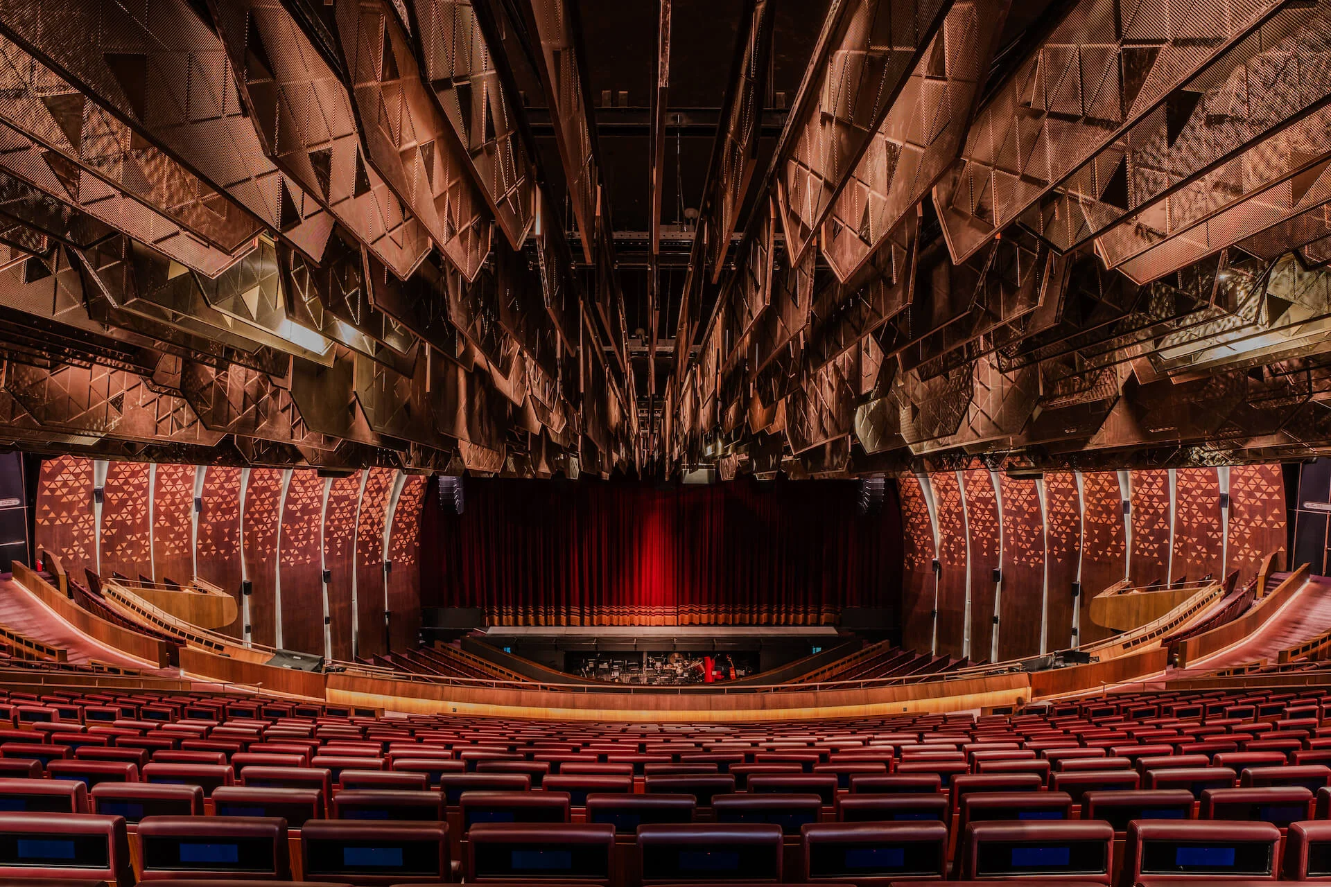 Sala del teatro dalla platea: poltrone rosse rivolte verso un sipario rosso sotto un soffitto di pannelli sfaccettati in rame – King Fahad Cultural Center, Riyadh, Arabia Saudita, 2024 – riqualificazione architettonica
