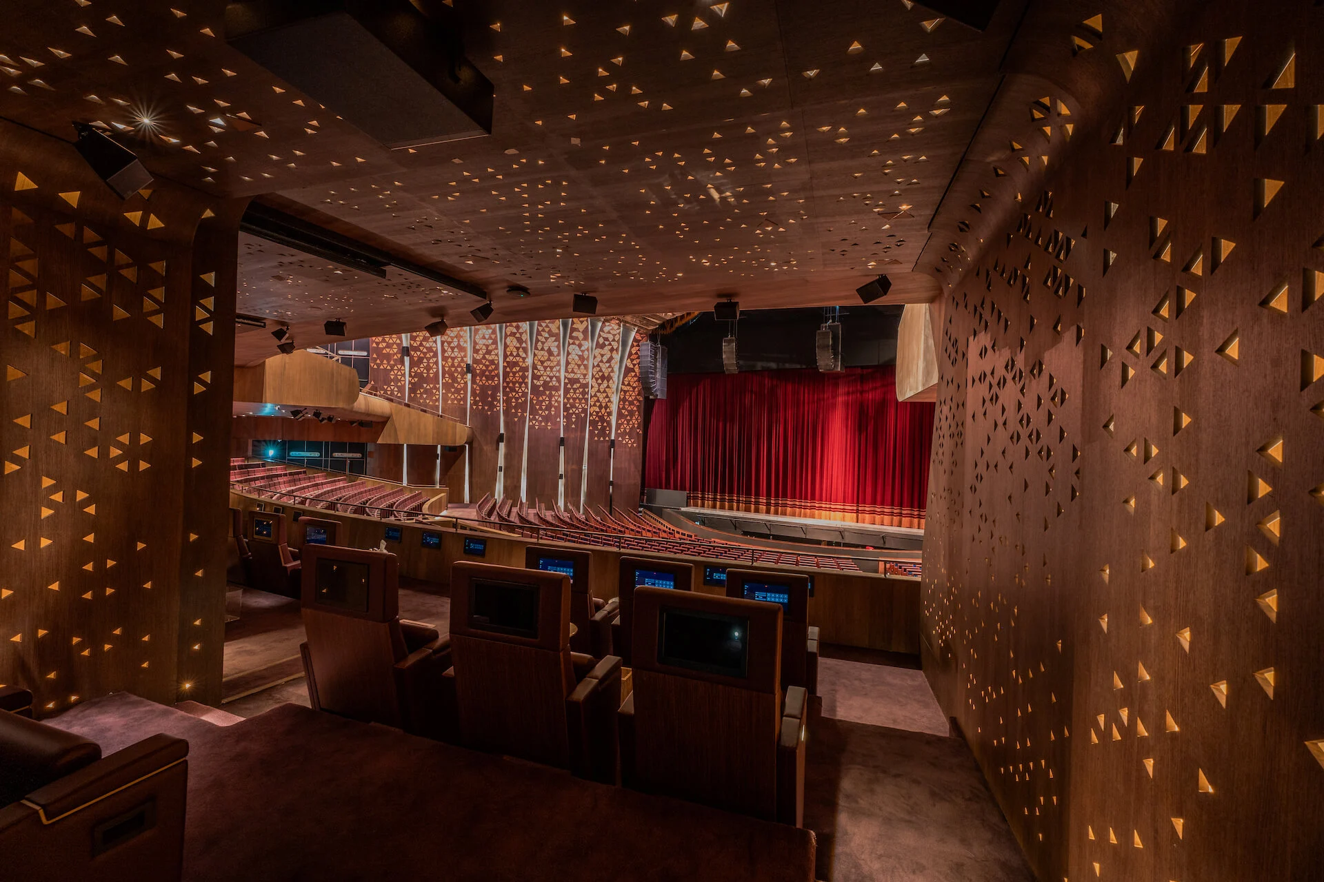 Vista da un palco attraverso pareti in legno forate da luci triangolari verso la scena e il sipario rosso – King Fahad Cultural Center, Riyadh, Arabia Saudita, 2024 – riqualificazione architettonica