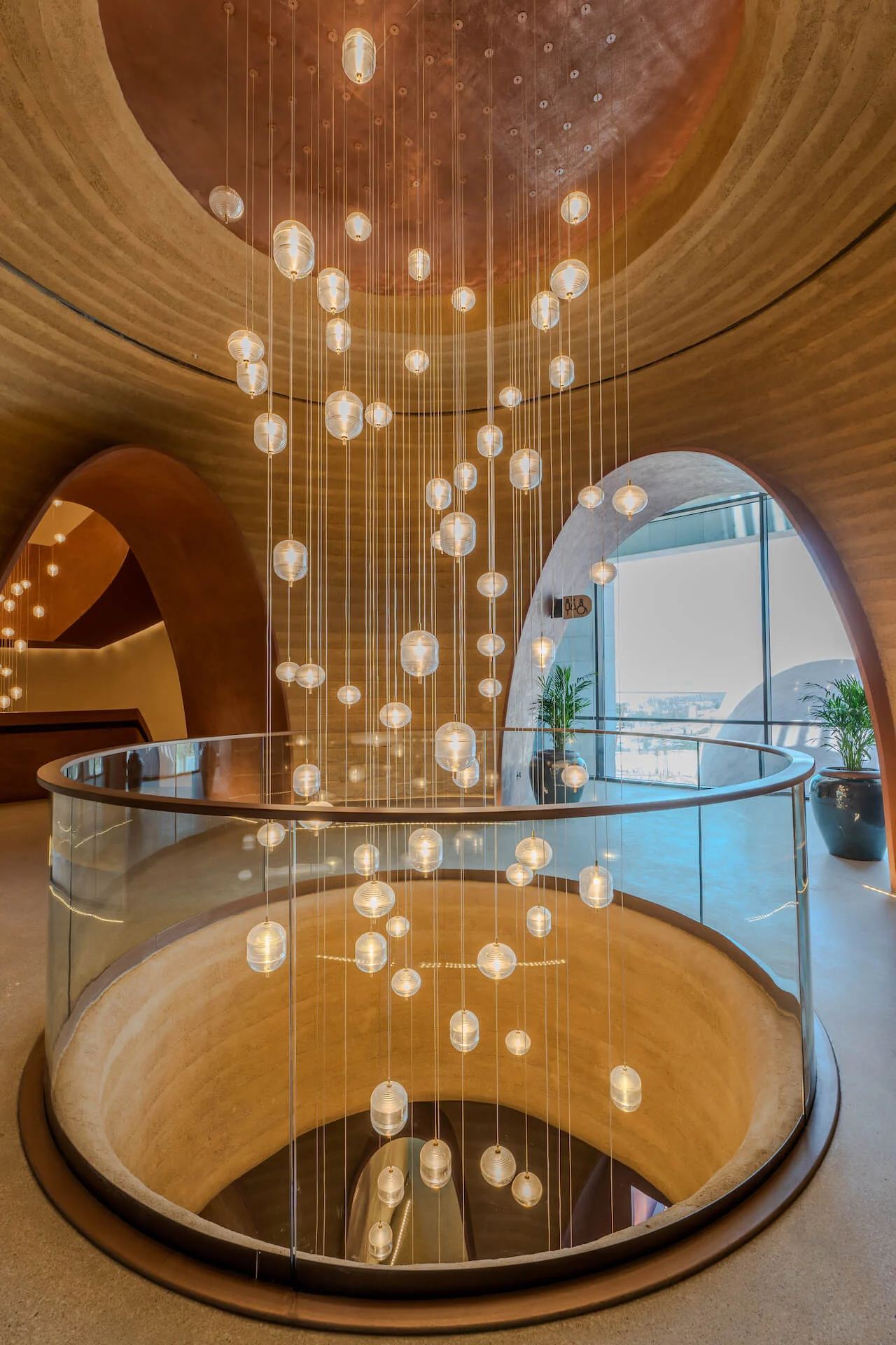 Vano scala circolare con parapetto in vetro e una cascata di lampade a sfera in vetro sotto una cupola in rame – King Fahad Cultural Center, Riyadh, Arabia Saudita, 2024 – riqualificazione architettonica