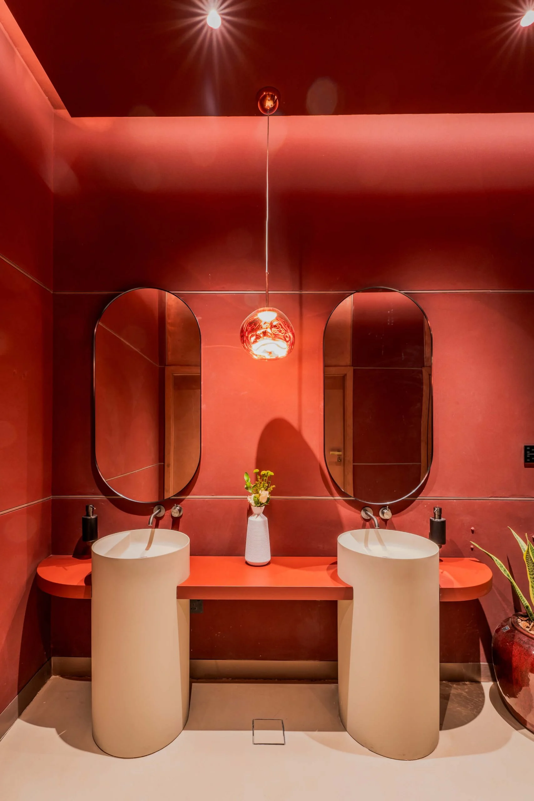 Vista frontale del bagno rosso: due lavabi bianchi a colonna sotto specchi ovali e una lampada a sospensione in rame – King Fahad Cultural Center, Riyadh, Arabia Saudita, 2024 – riqualificazione architettonica