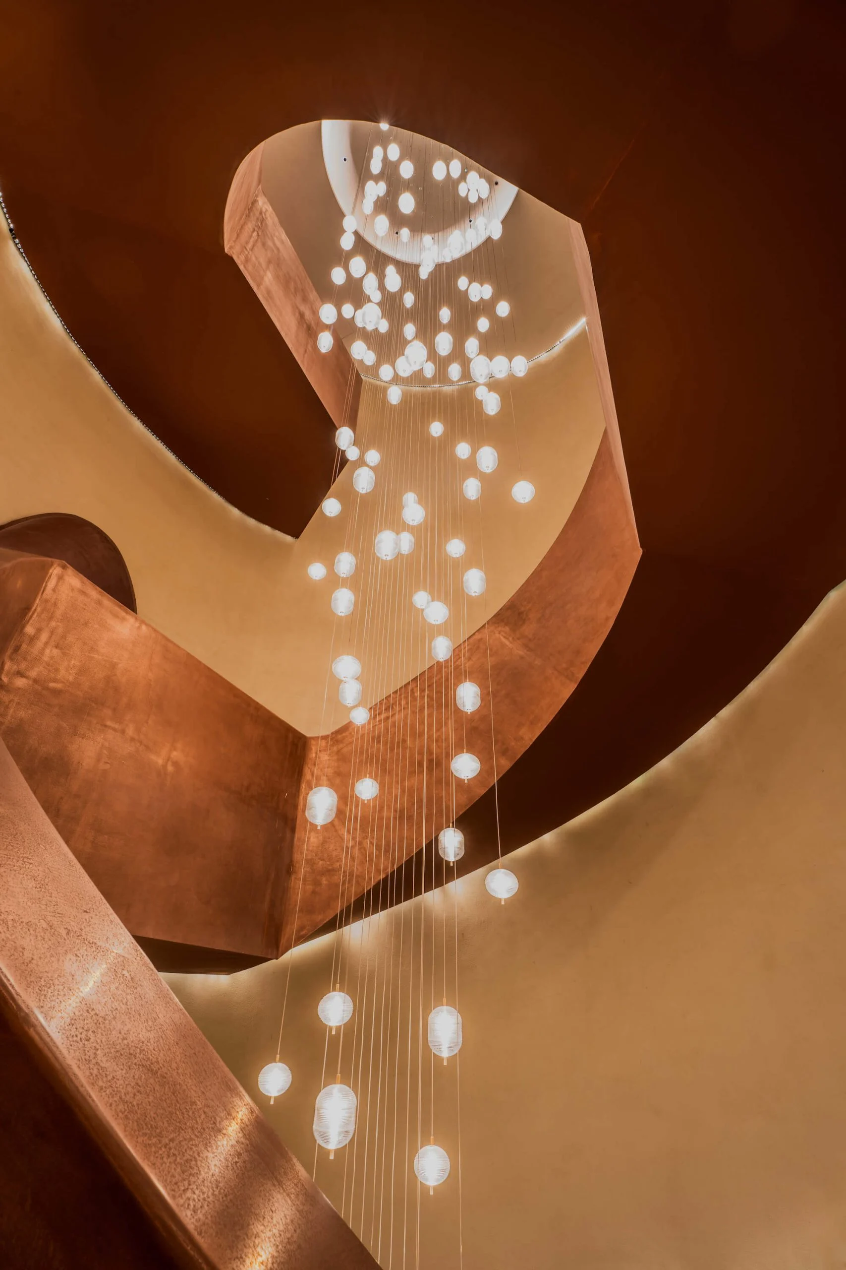 Vista dal basso della scala elicoidale in rame verso una cascata di lampade a sfera in vetro sospese – King Fahad Cultural Center, Riyadh, Arabia Saudita, 2024 – riqualificazione architettonica