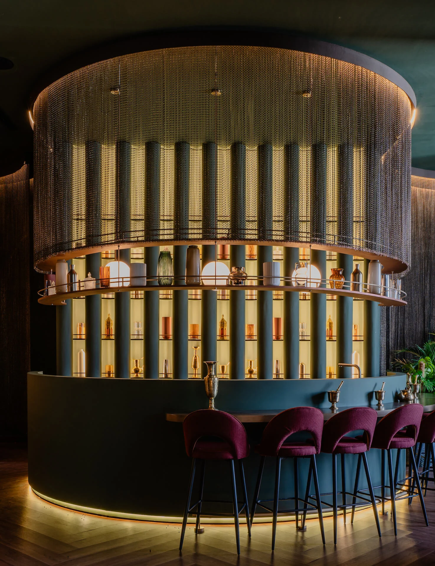 Bar circolare con bottiglie su mensole illuminate dietro una tenda di catene metalliche, e sgabelli in velluto bordeaux – King Fahad Cultural Center, Riyadh, Arabia Saudita, 2024 – riqualificazione architettonica