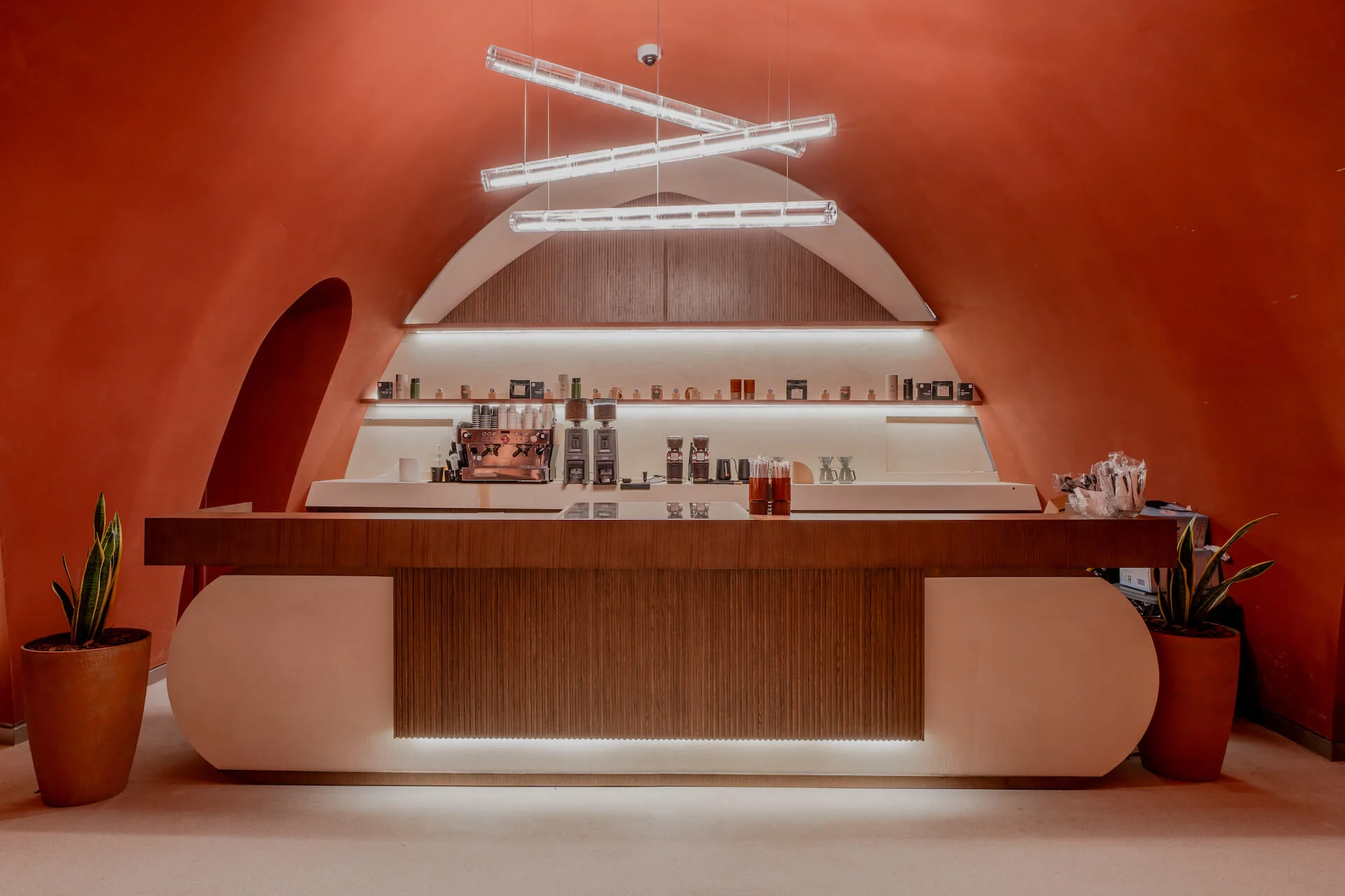 Caffetteria con bancone in legno e bianco sotto lampade lineari sospese, in una nicchia ad arco in terracotta – King Fahad Cultural Center, Riyadh, Arabia Saudita, 2024 – riqualificazione architettonica
