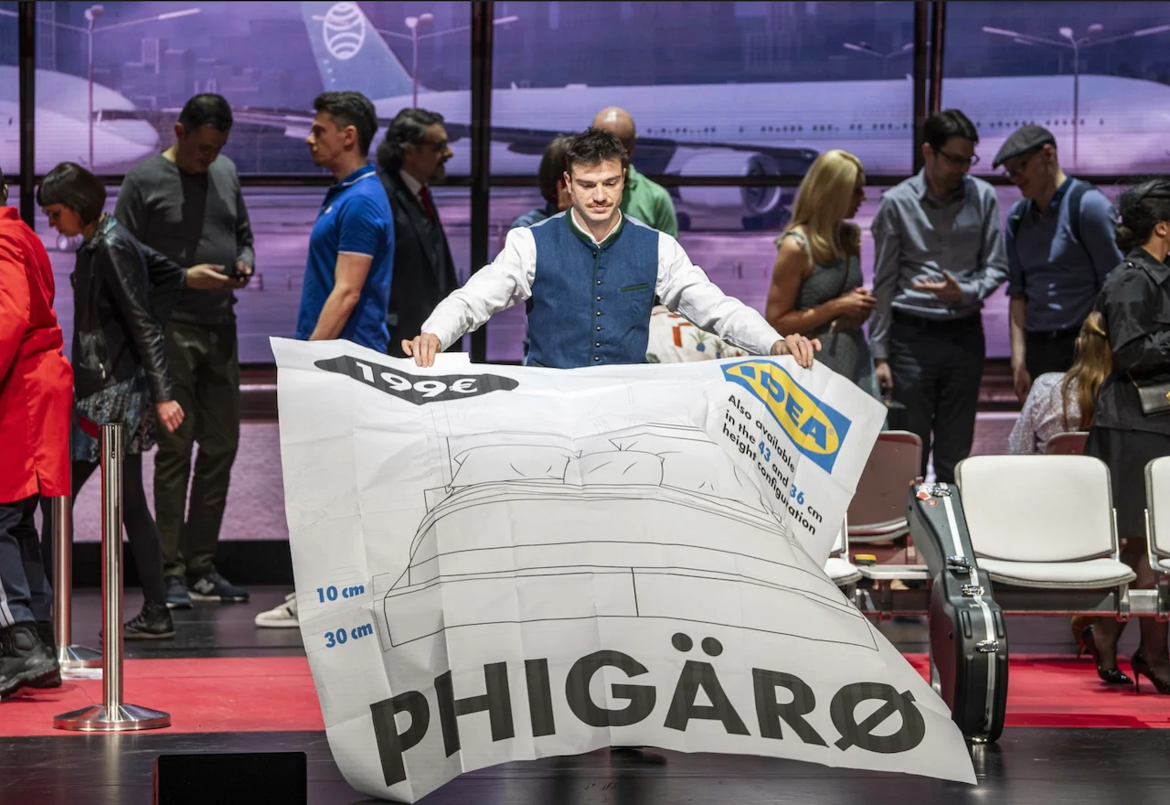 Cantante che solleva un gigantesco foglio di catalogo di mobili con la scritta Phigärø nel terminal dell'aeroporto – Une Folle Journée Mozart Gala, Salisburgo, Austria, 2024 – scenografia d'opera