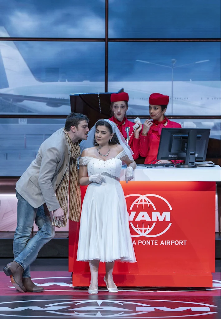 Sposa in bianco appoggiata al banco del check-in WAM mentre un uomo le sussurra, assistenti di volo dietro – Une Folle Journée Mozart Gala, Salisburgo, Austria, 2024 – scenografia d'opera