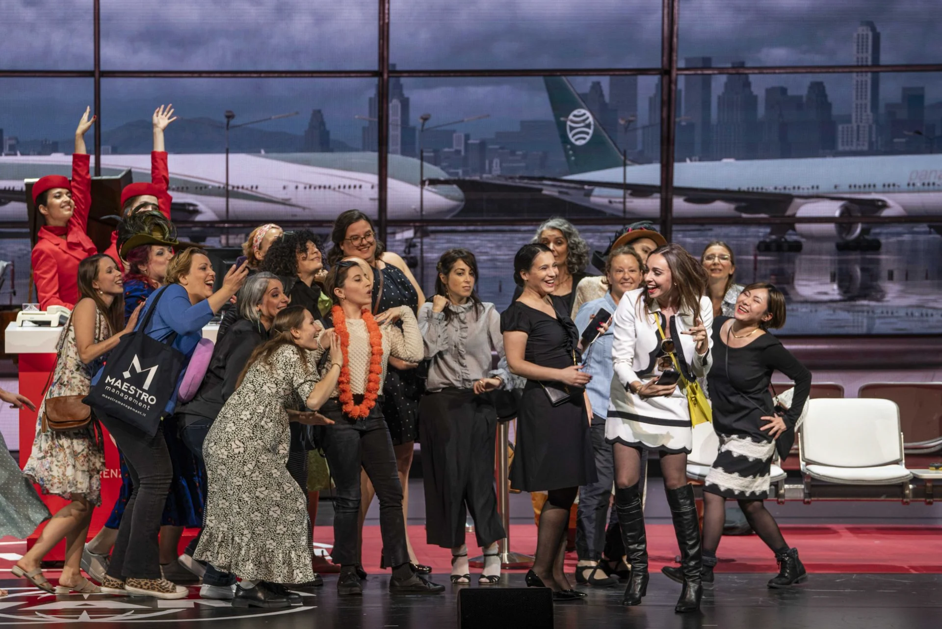 Gruppo di donne nel terminal raccolte intorno a una cantante in abito bianco e nero – Une Folle Journée Mozart Gala, Salisburgo, Austria, 2024 – scenografia d'opera