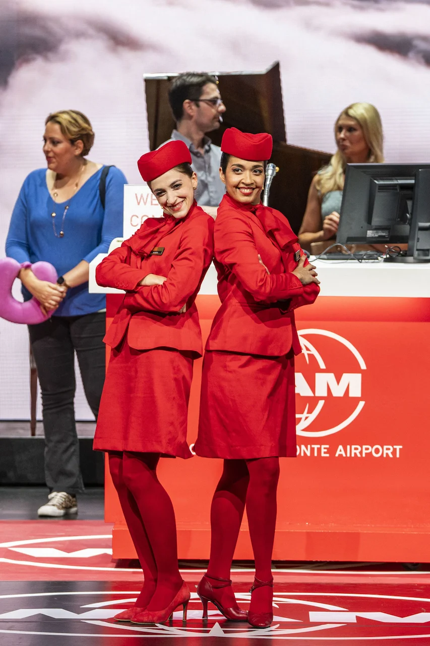 Due artisti in divisa rossa da assistente di volo in posa schiena contro schiena al banco WAM dell'aeroporto – Une Folle Journée Mozart Gala, Salisburgo, Austria, 2024 – scenografia d'opera