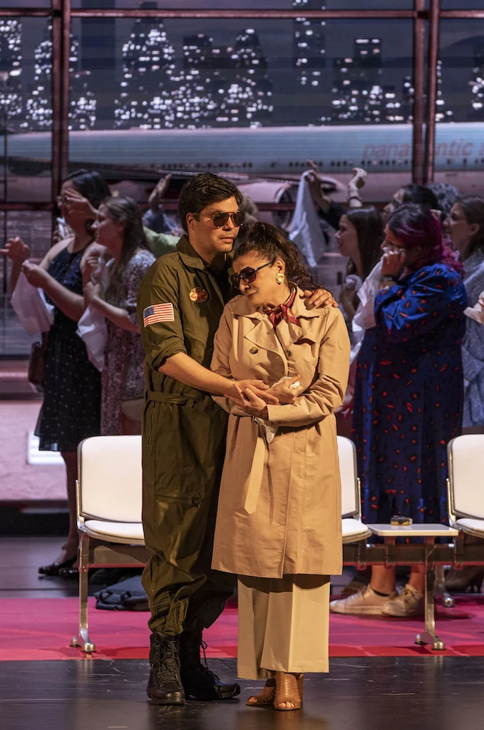 Coppia abbracciata, in tuta da pilota e trench, tra i passeggeri nel terminal – Une Folle Journée Mozart Gala, Salisburgo, Austria, 2024 – scenografia d'opera