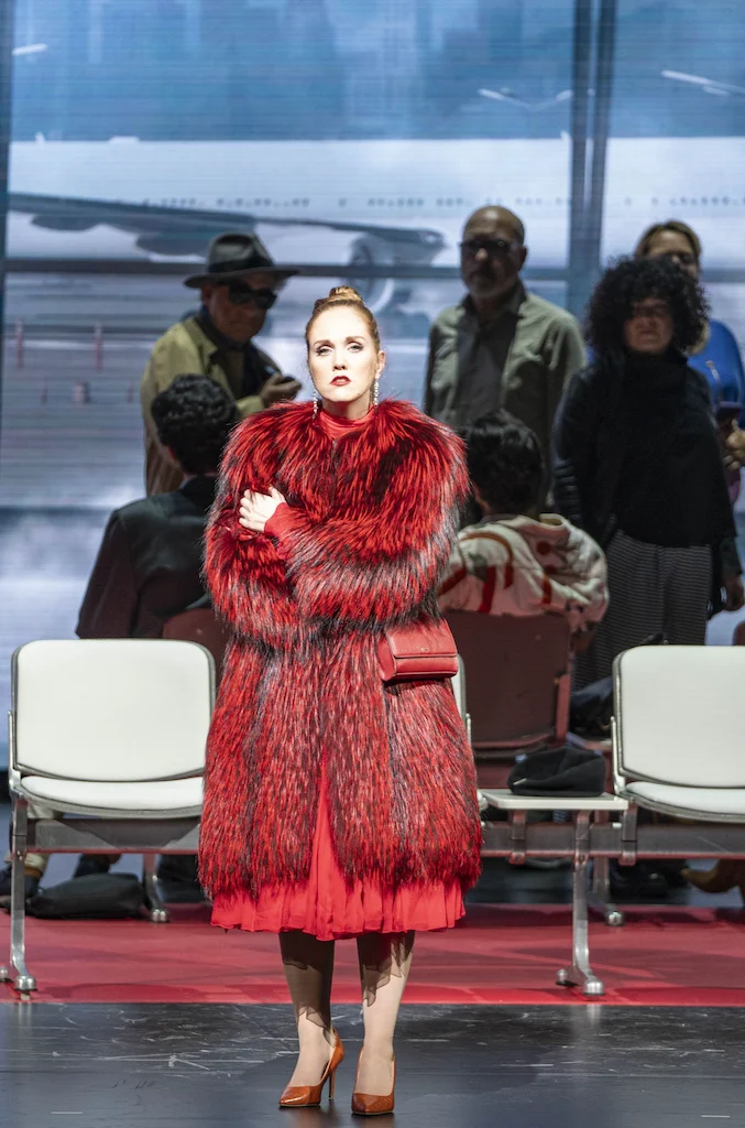Cantante in pelliccia rossa in piedi davanti a file di sedute dell'aeroporto e passeggeri – Une Folle Journée Mozart Gala, Salisburgo, Austria, 2024 – scenografia d'opera