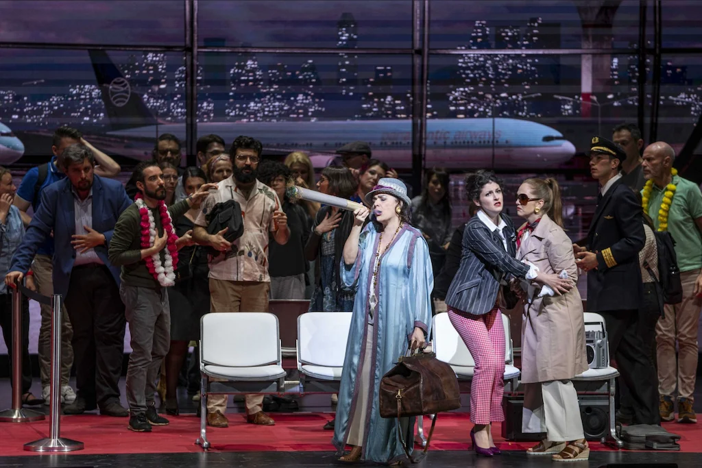 Cantante in vestaglia blu e cappello con una borsa da viaggio, circondato da passeggeri nel terminal di notte – Une Folle Journée Mozart Gala, Salisburgo, Austria, 2024 – scenografia d'opera