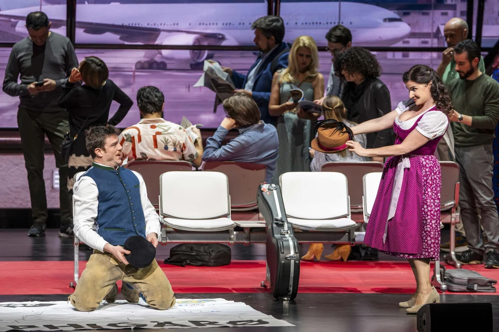 Cantante in gilet inginocchiato davanti a una cantante in dirndl rosa tra le sedute dell'aeroporto – Une Folle Journée Mozart Gala, Salisburgo, Austria, 2024 – scenografia d'opera