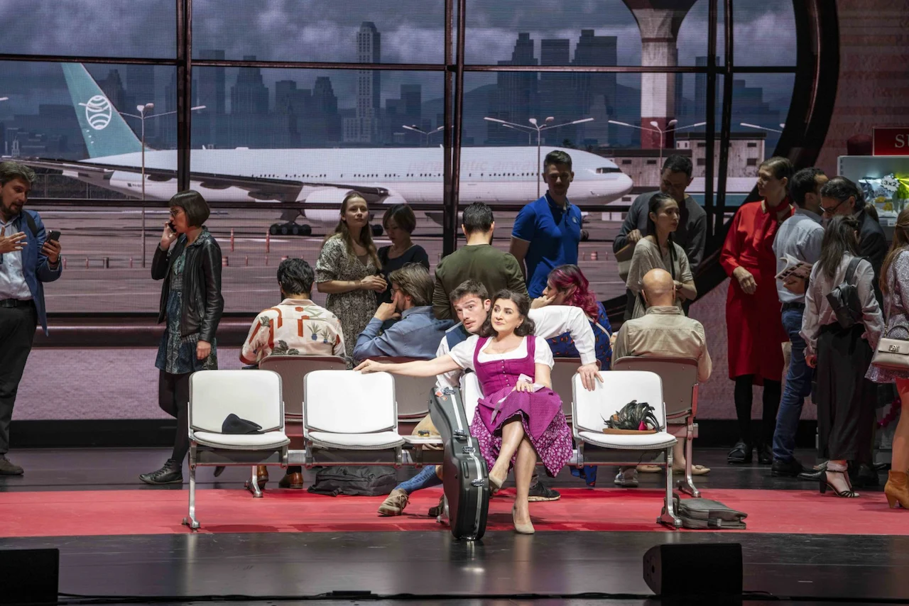 Cantante in abito rosa in posa sulle sedute dell'aeroporto con la custodia di un violoncello, passeggeri e un aereo dietro – Une Folle Journée Mozart Gala, Salisburgo, Austria, 2024 – scenografia d'opera