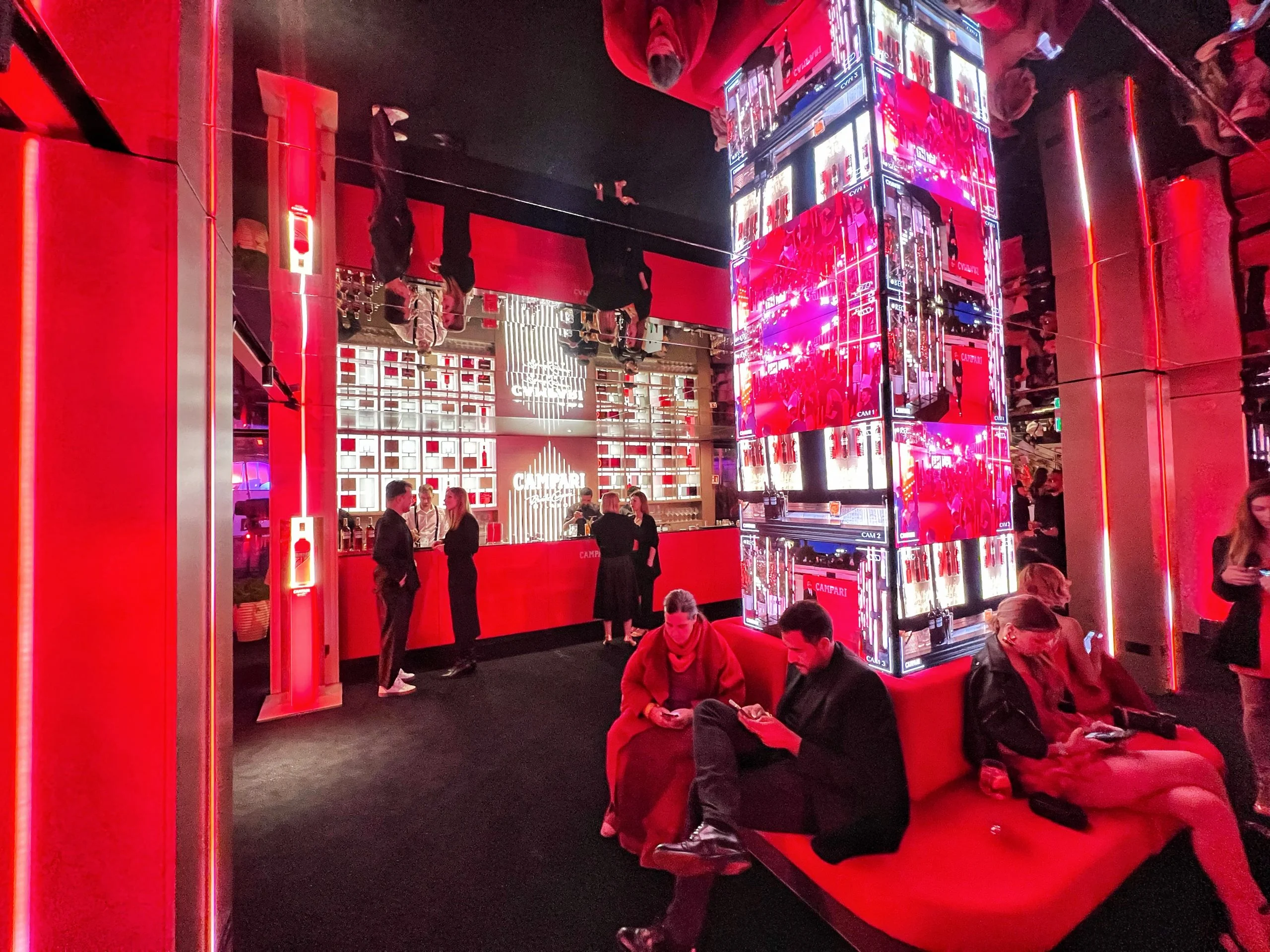 Lounge illuminata di rosso di notte con ospiti sui divani rossi, soffitto a specchio e una colonna rivestita di schermi video – Campari al Festival di Cannes 2024, Francia – architettura per eventi