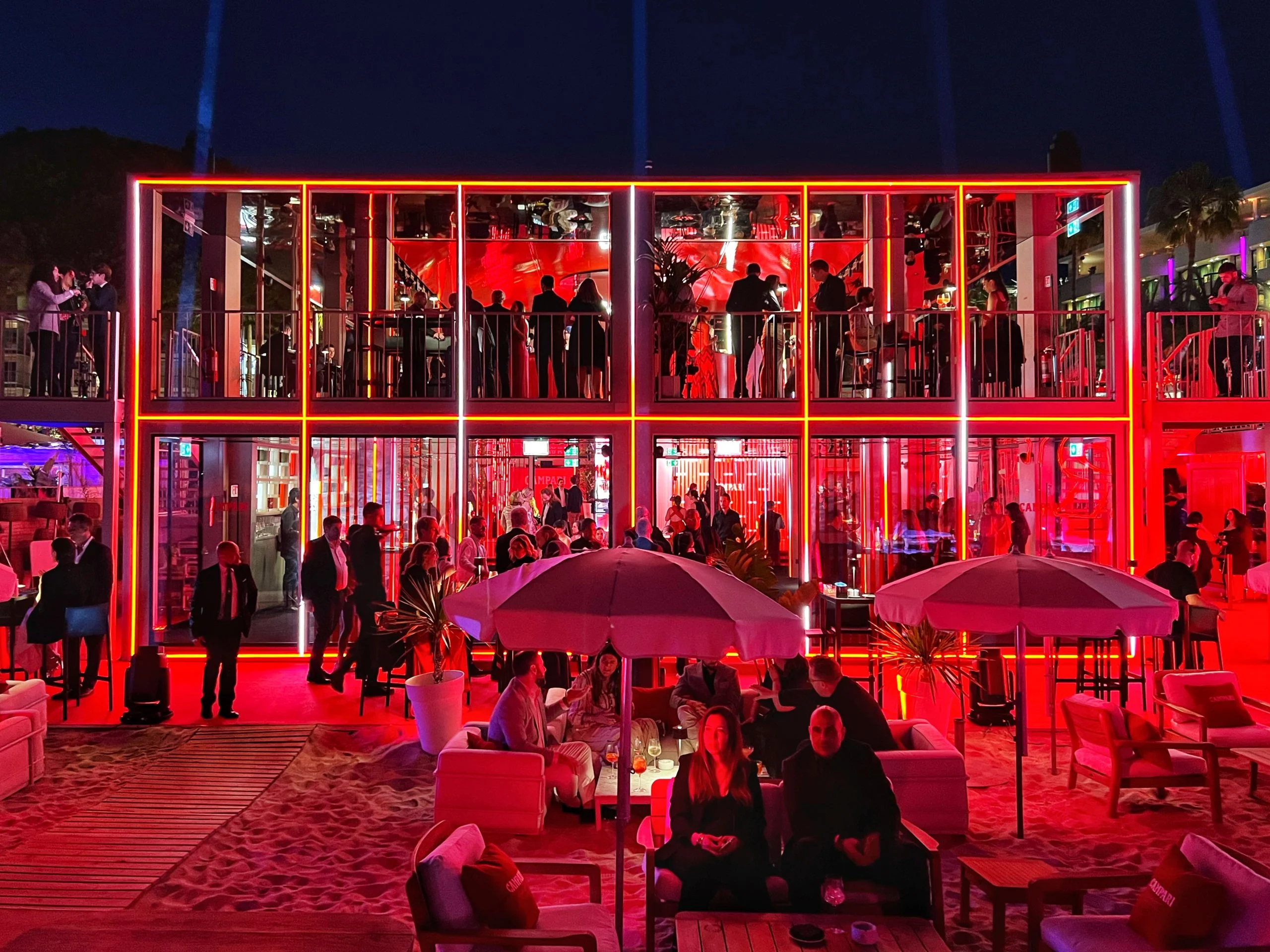 Facciata del padiglione accesa di rosso di notte, ospiti su entrambi i piani e sotto gli ombrelloni sulla sabbia – Campari al Festival di Cannes 2024, Francia – architettura per eventi