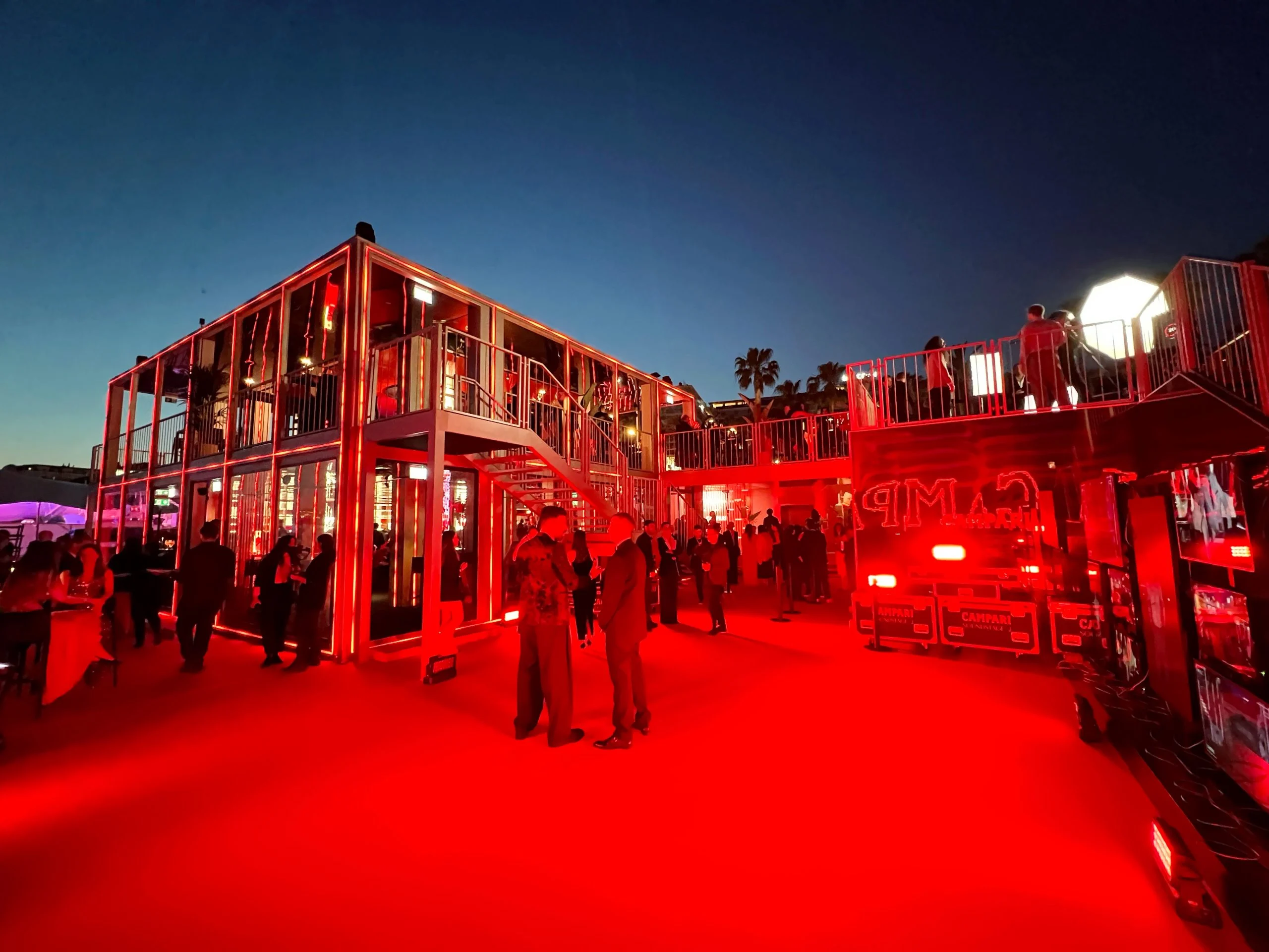 Piazzale illuminato di rosso al tramonto con il padiglione in vetro a due piani, le scale esterne e una terrazza rossa – Campari al Festival di Cannes 2024, Francia – architettura per eventi