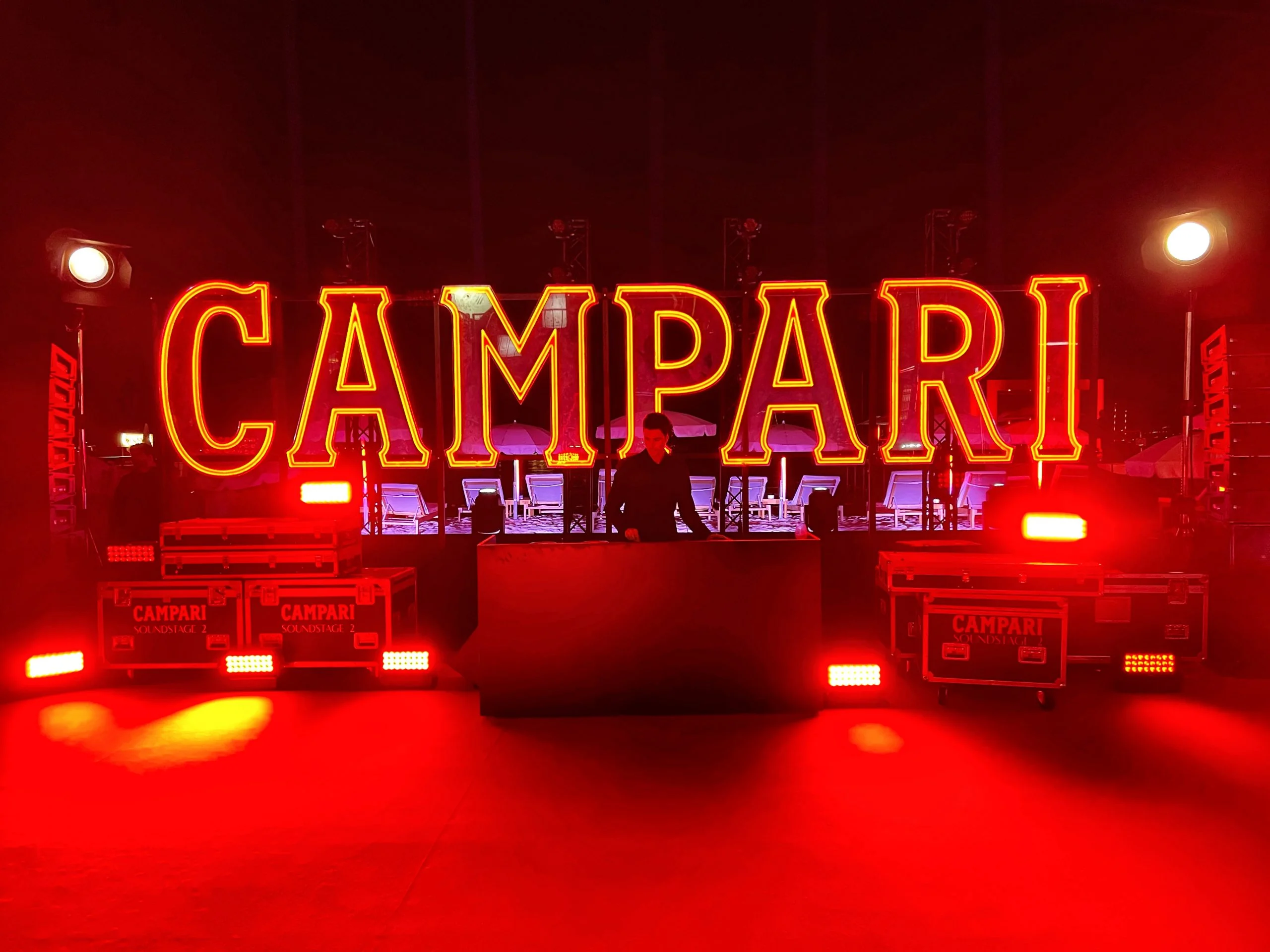 Gigantesche lettere Campari illuminate sopra una postazione DJ, tra flight case e luci rosse – Campari al Festival di Cannes 2024, Francia – architettura per eventi
