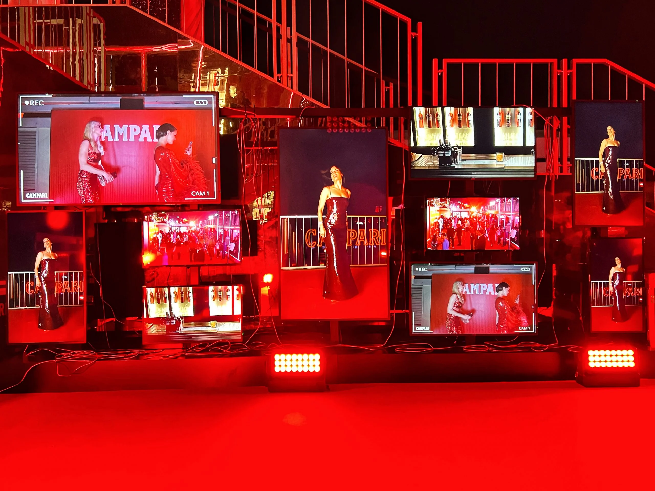 Parete di schermi con le riprese dal vivo degli ospiti in posa davanti a un fondale Campari, scale illuminate di rosso dietro – Campari al Festival di Cannes 2024, Francia – architettura per eventi