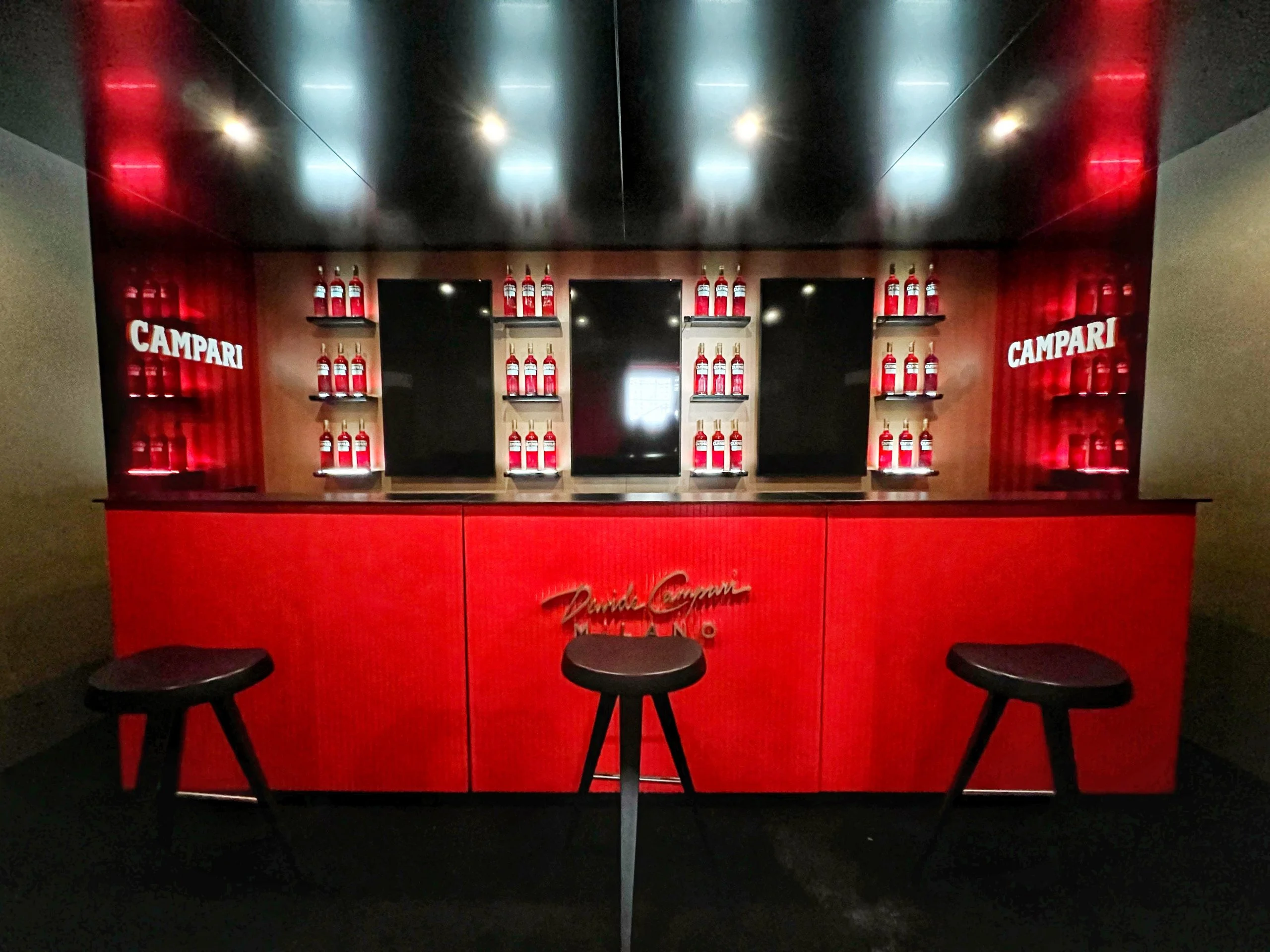 Bancone bar rosso con la scritta Paolo Campari Milano, tre sgabelli neri e scaffali di bottiglie Campari – Campari al Festival di Cannes 2024, Francia – architettura per eventi