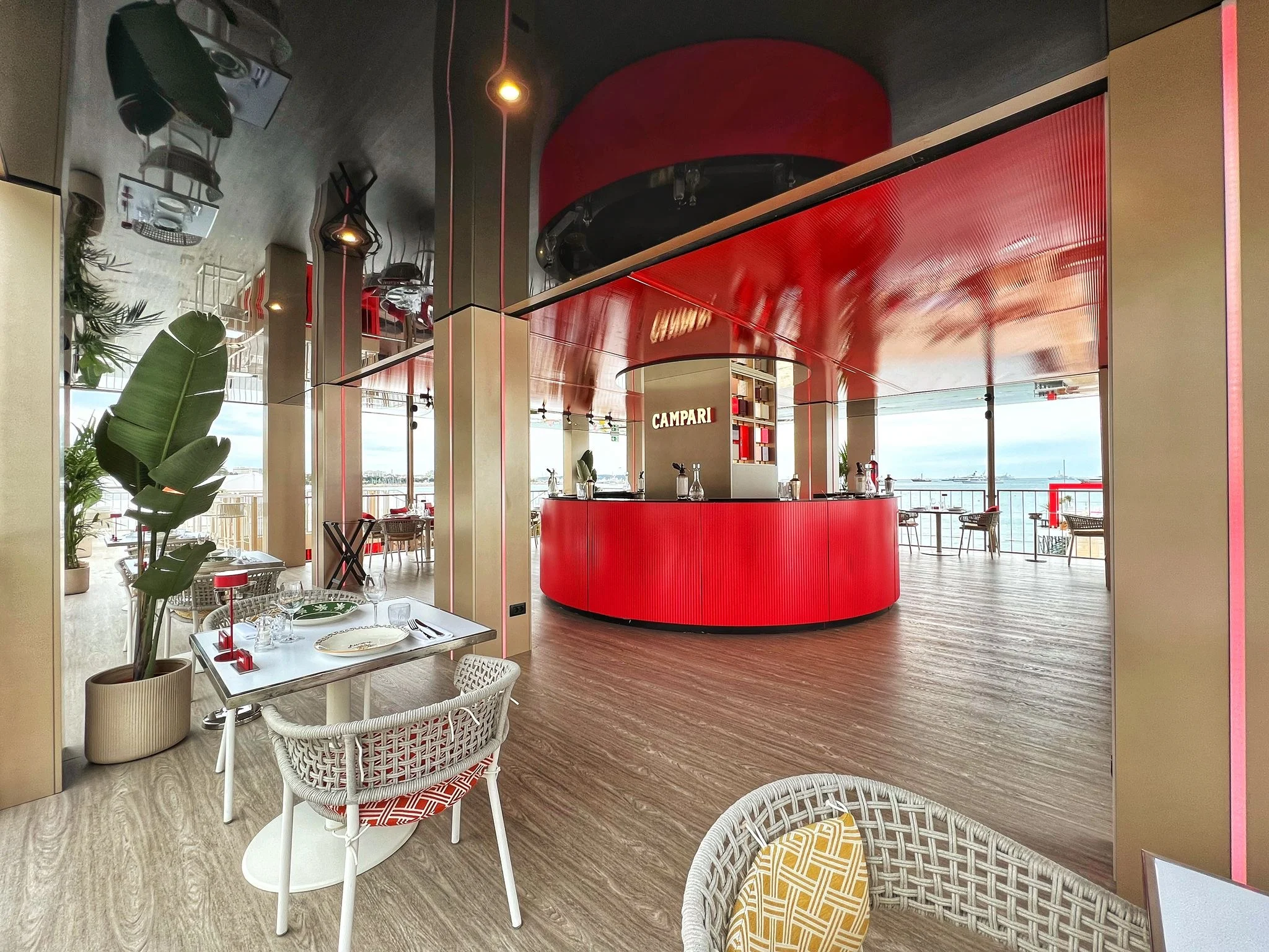 Zona pranzo in terrazza con sedie in vimini e piante, verso il bar rotondo rosso Campari – Campari al Festival di Cannes 2024, Francia – architettura per eventi