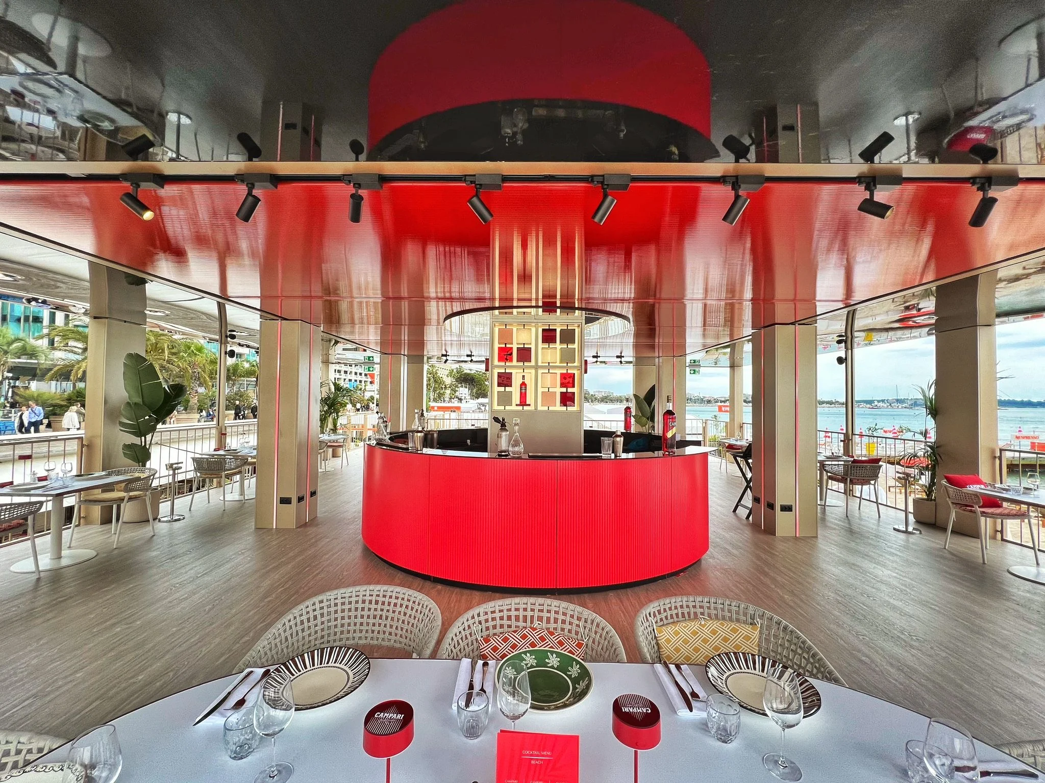Ristorante in terrazza con bar rotondo rosso sotto un soffitto rosso lucido, tavoli apparecchiati in primo piano – Campari al Festival di Cannes 2024, Francia – architettura per eventi