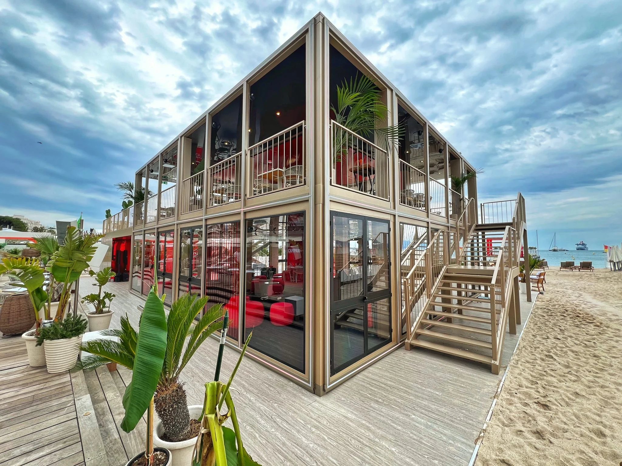 Padiglione a due piani in vetro e oro champagne sulla spiaggia di Cannes, con scale in legno e palme in vaso – Campari al Festival di Cannes 2024, Francia – architettura per eventi