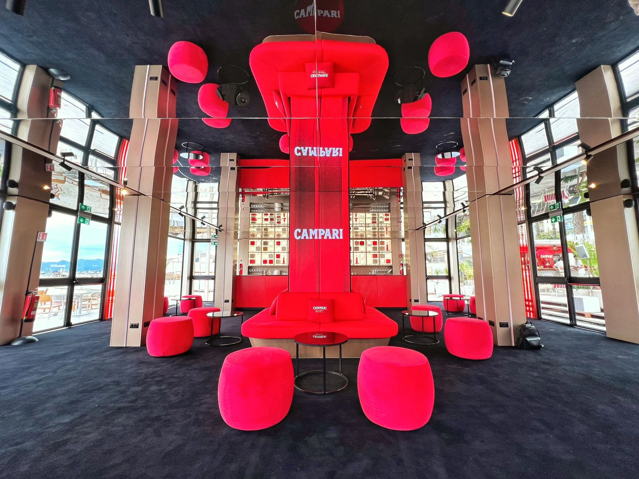 Lounge buia con una colonna rossa centrale, pouf e divani rossi e cuscini rossi appesi al soffitto a specchio – Campari al Festival di Cannes 2024, Francia – architettura per eventi