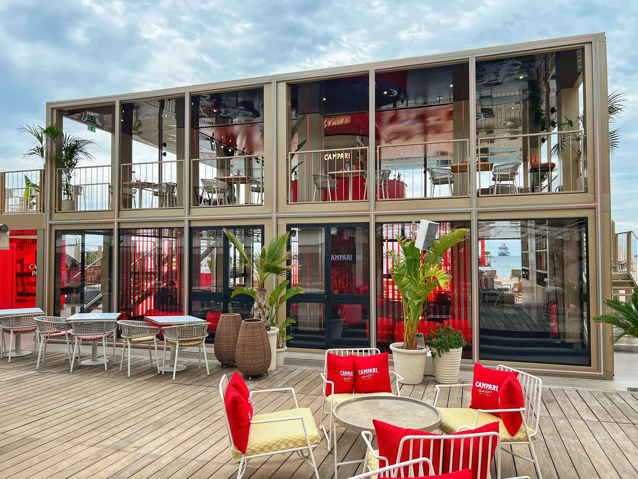 Facciata vetrata del padiglione Campari con interni rossi, sedute sulla terrazza con cuscini rossi Campari davanti – Campari al Festival di Cannes 2024, Francia – architettura per eventi