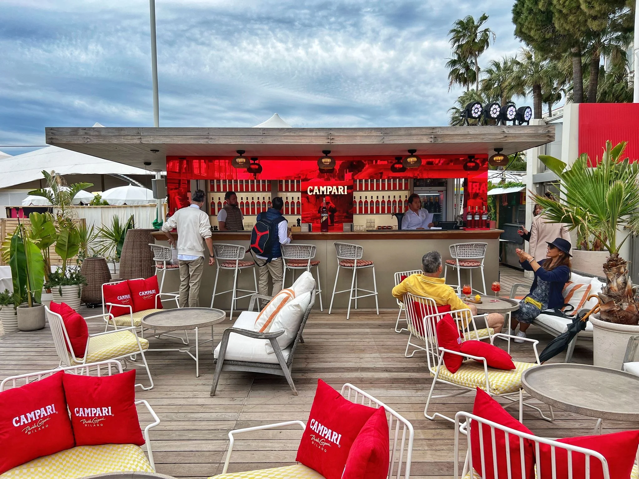 Bar sulla spiaggia all'aperto con fondale rosso Campari, sgabelli e sedie bianche con cuscini rossi – Campari al Festival di Cannes 2024, Francia – architettura per eventi