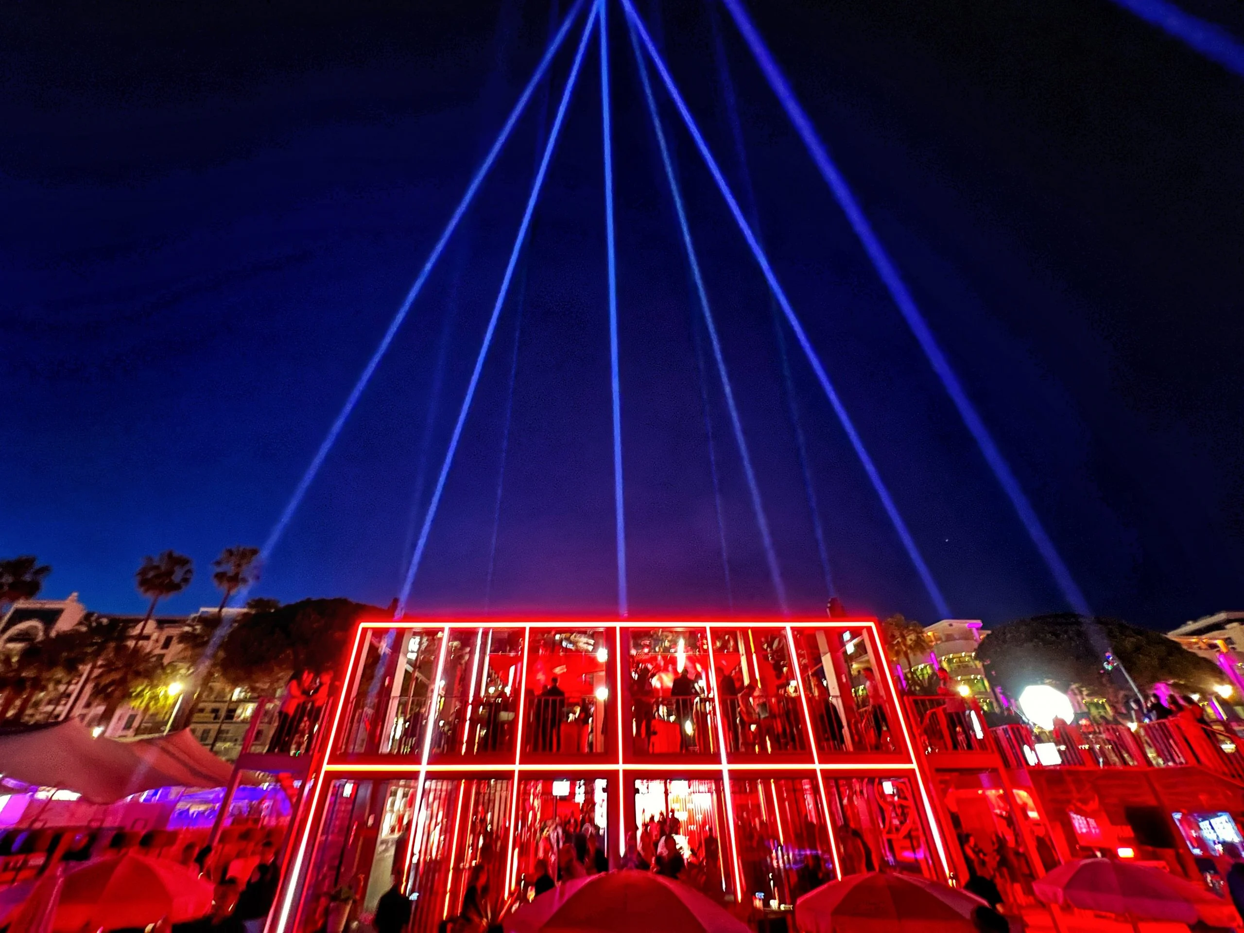 Vista notturna del padiglione profilato di luce rossa, con fasci di fari a ventaglio nel cielo sopra – Campari al Festival di Cannes 2024, Francia – architettura per eventi