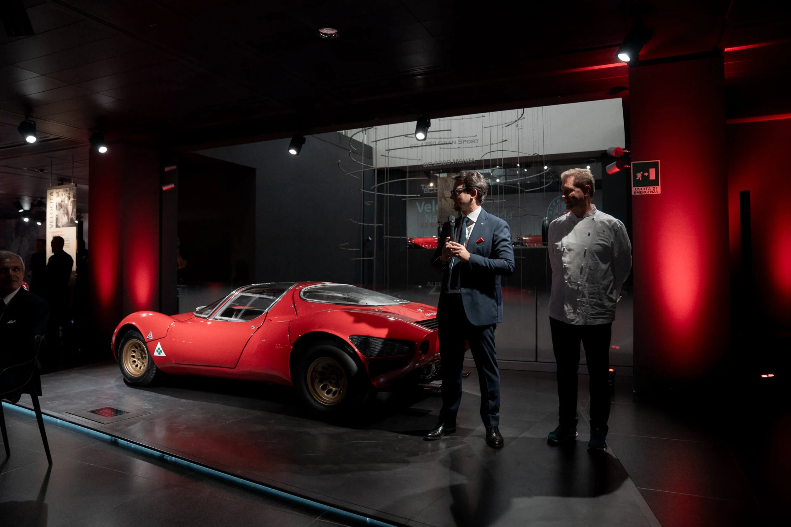 Due relatori accanto all'originale Alfa Romeo 33 Stradale rossa in una sala del museo illuminata di rosso – Presentazione Alfa Romeo 33 Stradale, Arese, Italia, 2023 – progettazione di eventi