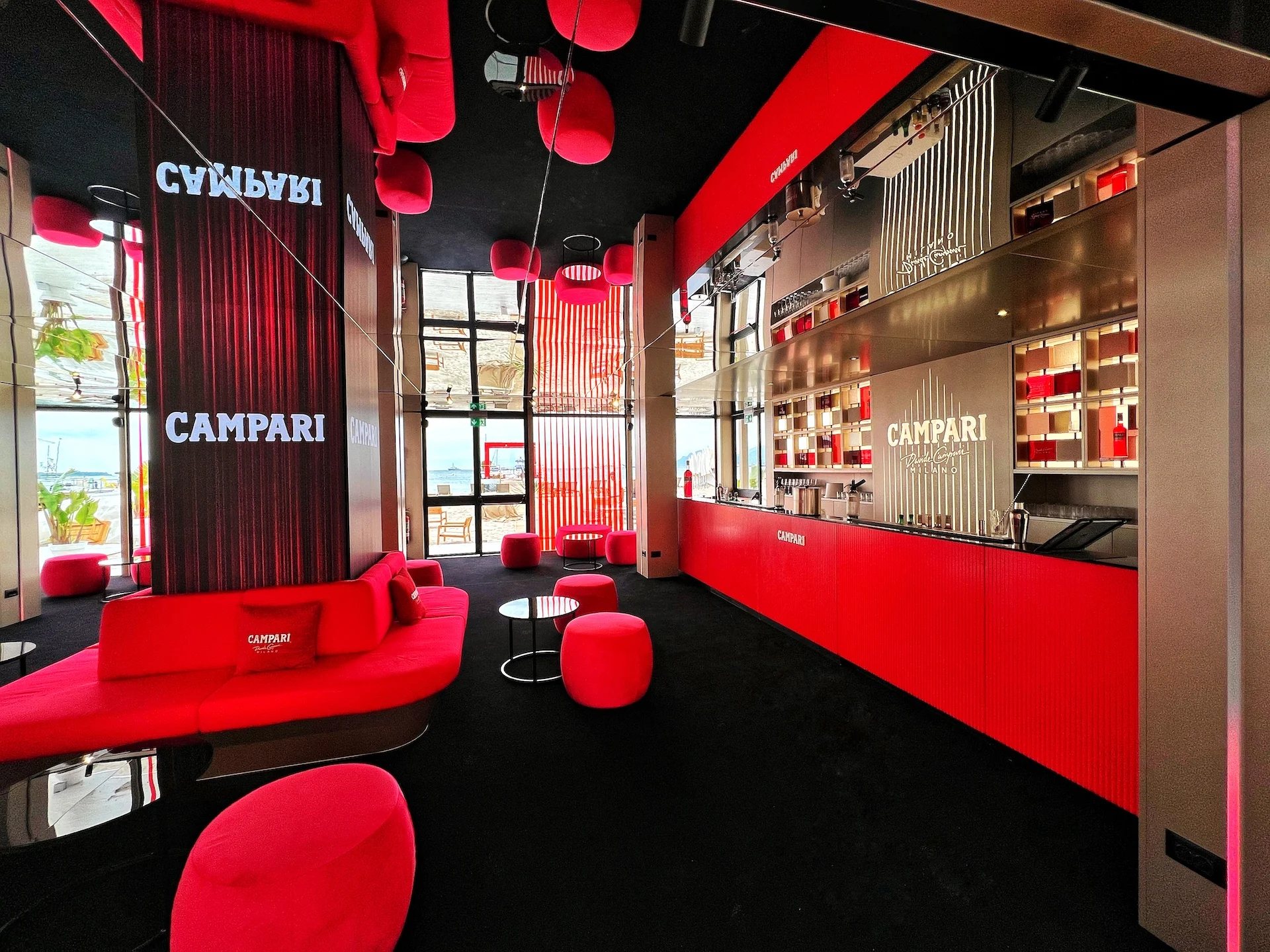Lounge al piano terra con divani e pouf rossi, una colonna con il marchio Campari e un lungo bancone bar rosso – Campari al Festival di Cannes 2024, Francia – architettura per eventi
