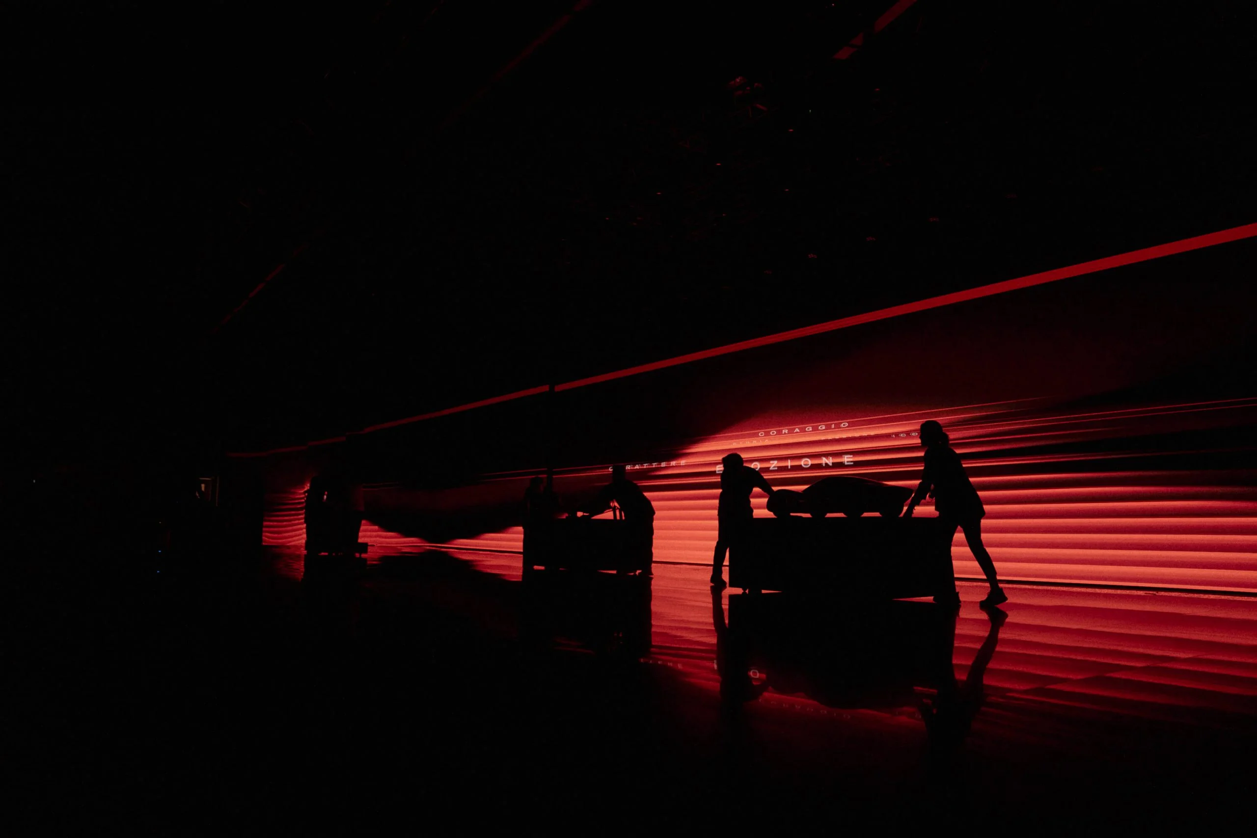 Sagome di tecnici che spingono auto coperte su un palco illuminato di rosso davanti a una lunga parete LED – Presentazione Alfa Romeo 33 Stradale, Arese, Italia, 2023 – progettazione di eventi