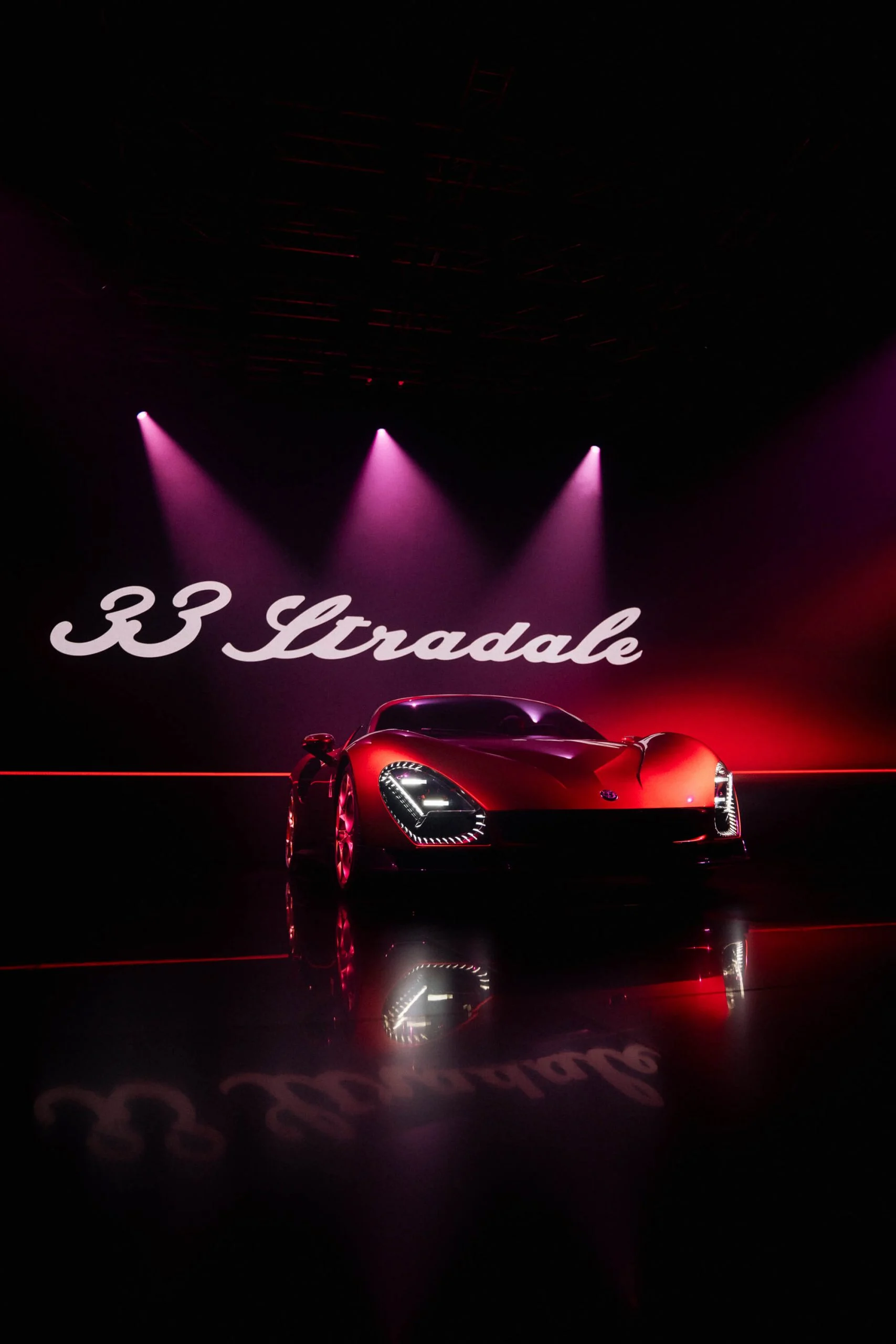 33 Stradale rossa sotto tre riflettori rosa, con il nome del modello in corsivo bianco sulla parete dietro – Presentazione Alfa Romeo 33 Stradale, Arese, Italia, 2023 – progettazione di eventi