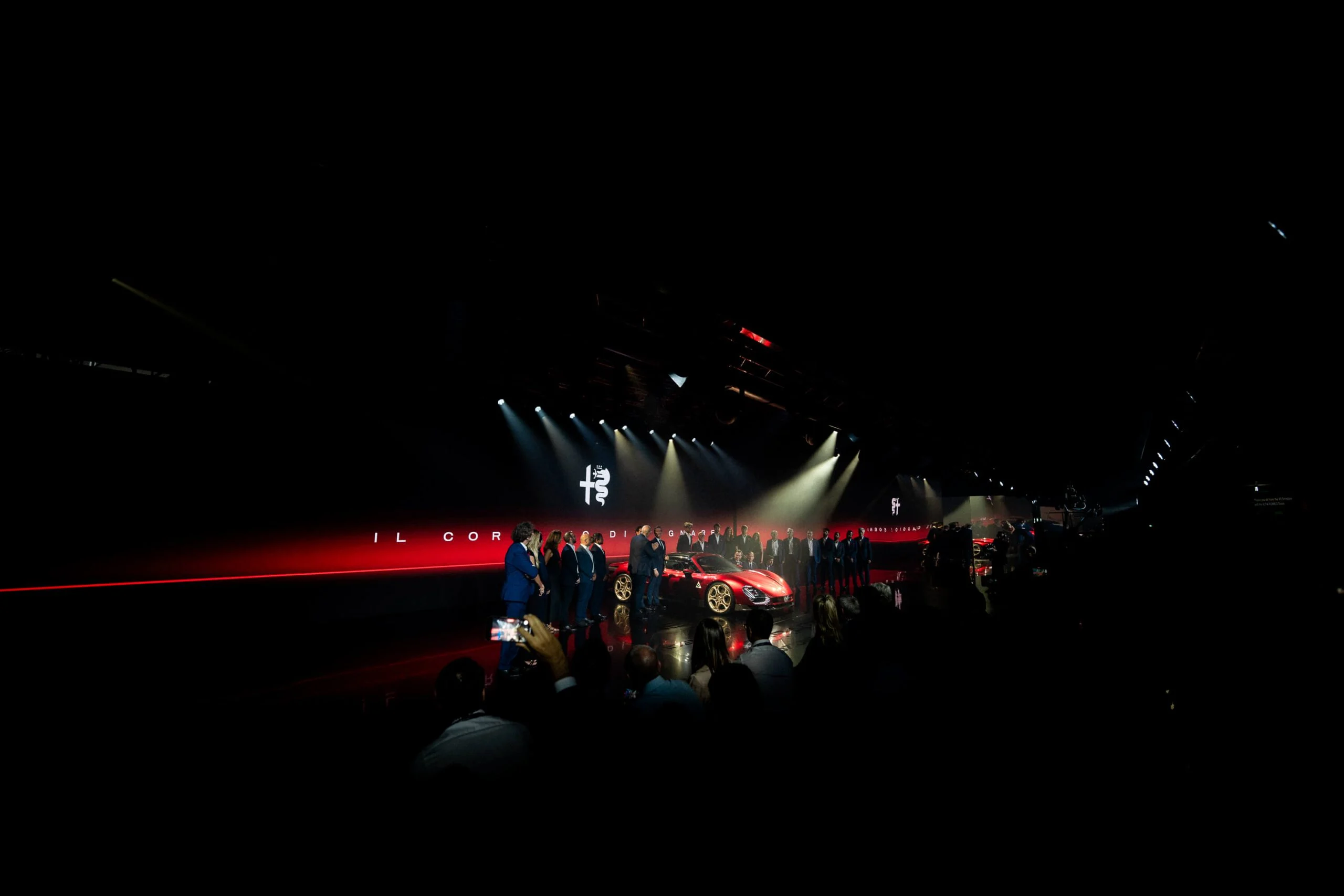Vista d'insieme della sala buia: ospiti che filmano l'auto sul palco sotto il logo Alfa Romeo – Presentazione Alfa Romeo 33 Stradale, Arese, Italia, 2023 – progettazione di eventi