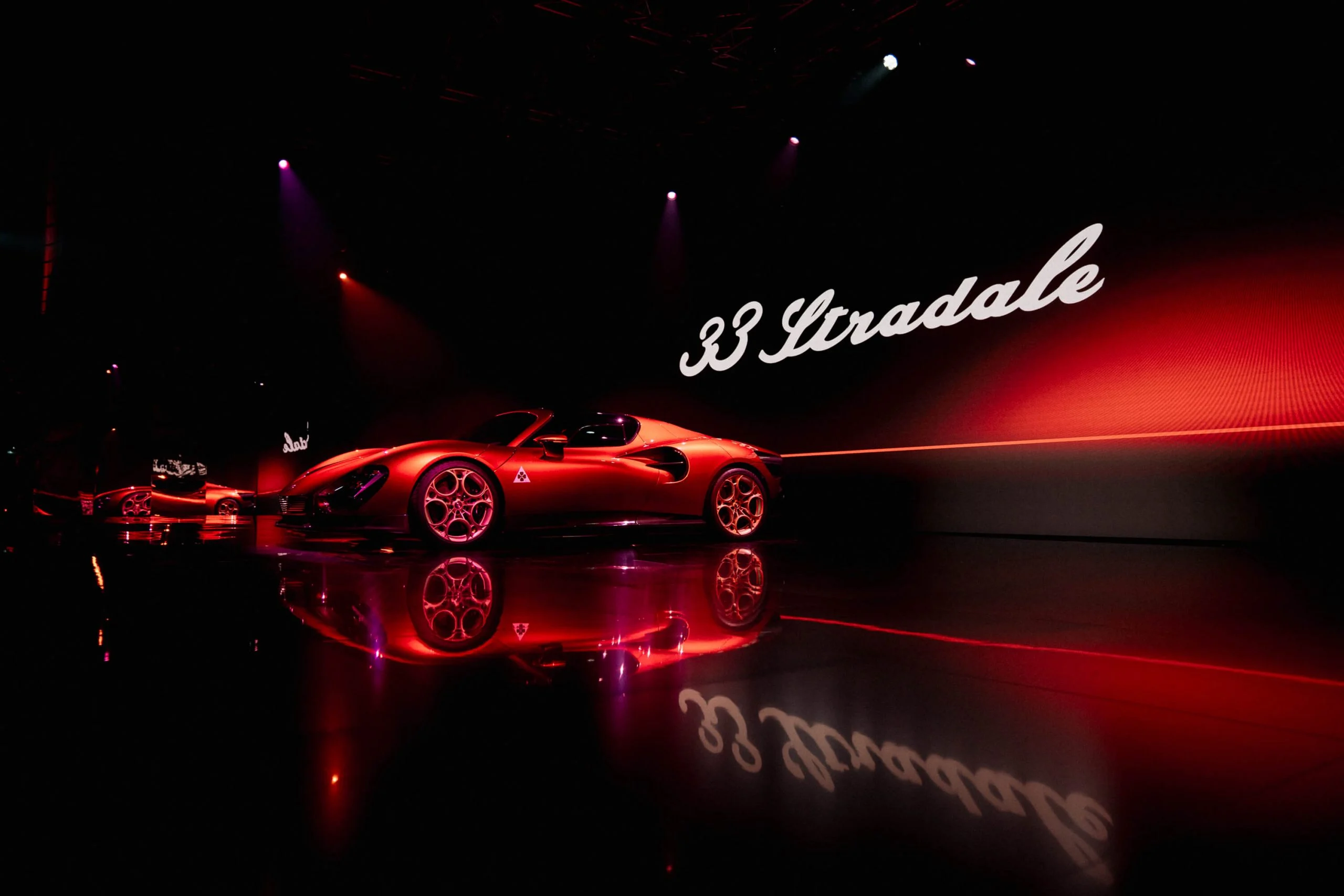 33 Stradale rossa con il suo nome in corsivo bianco sulla parete, riflessa sul pavimento nero – Presentazione Alfa Romeo 33 Stradale, Arese, Italia, 2023 – progettazione di eventi