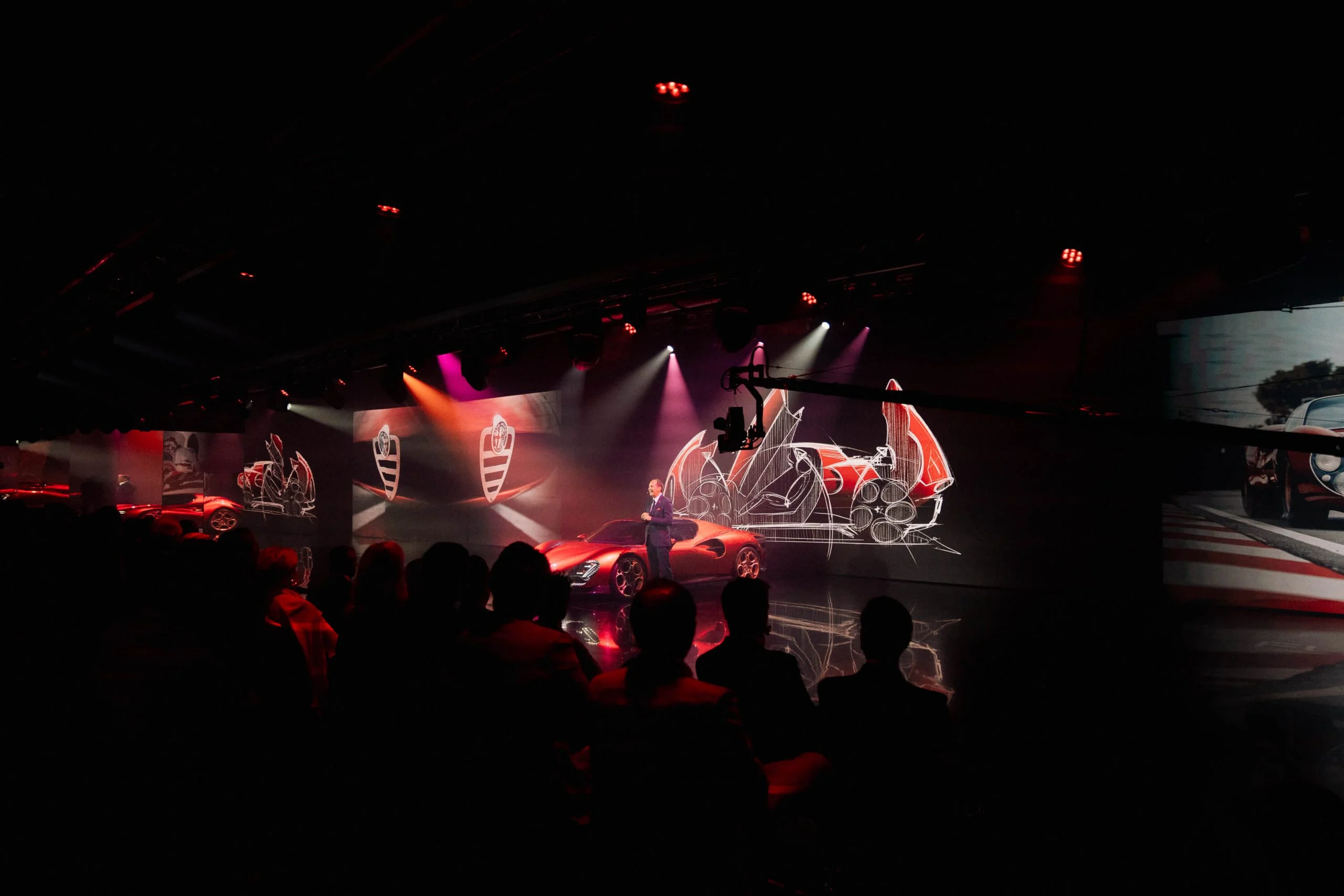 Presentatore sul palco accanto all'auto rossa, con schizzi proiettati della 33 Stradale dietro – Presentazione Alfa Romeo 33 Stradale, Arese, Italia, 2023 – progettazione di eventi