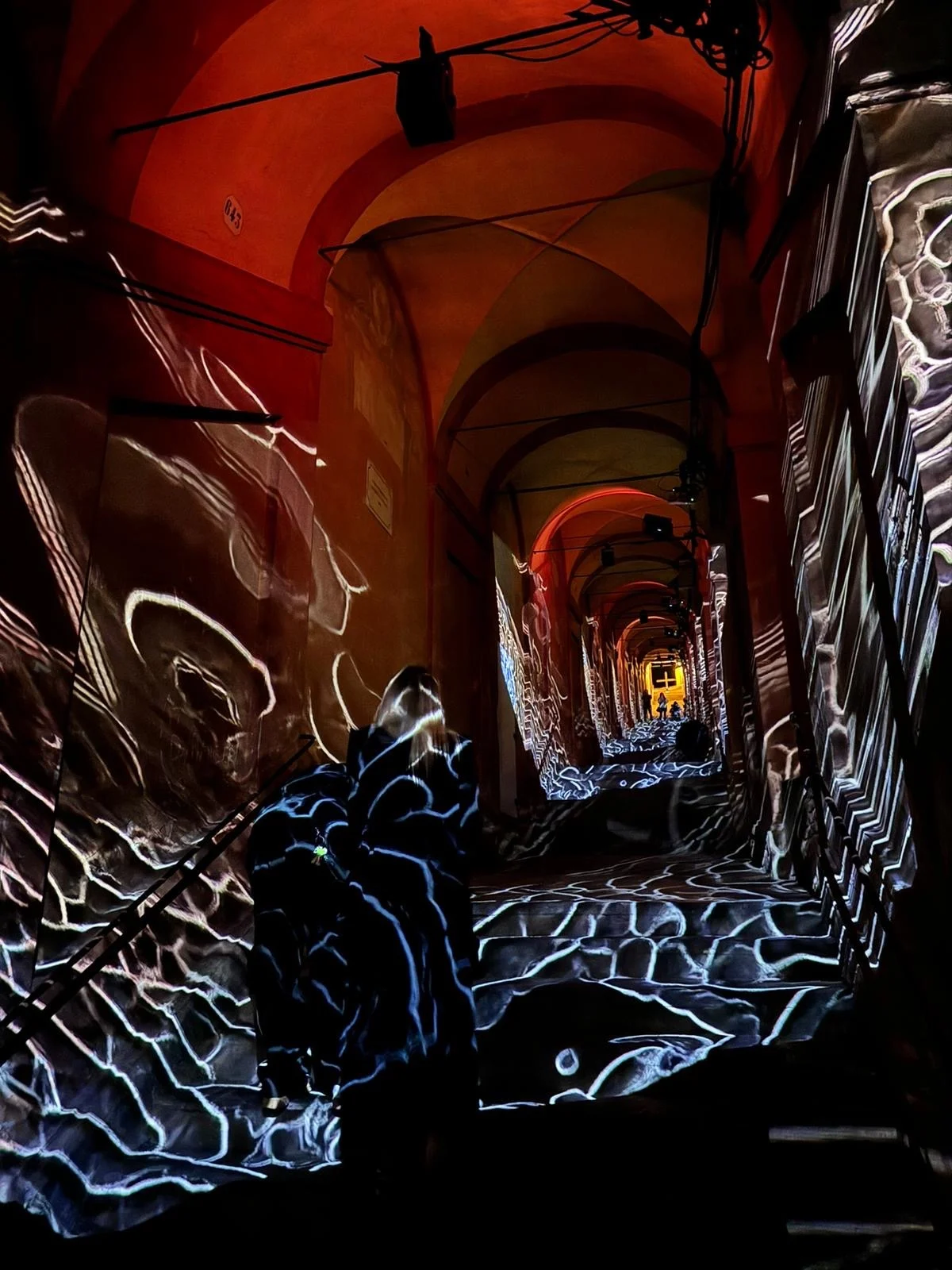 Proiezioni bianche a onde su pareti e gradini del portico sotto volte illuminate di rosso, un visitatore nella luce – Cesare Cremonini Festival dei Portici, Bologna, Italia, 2024 – progettazione di eventi