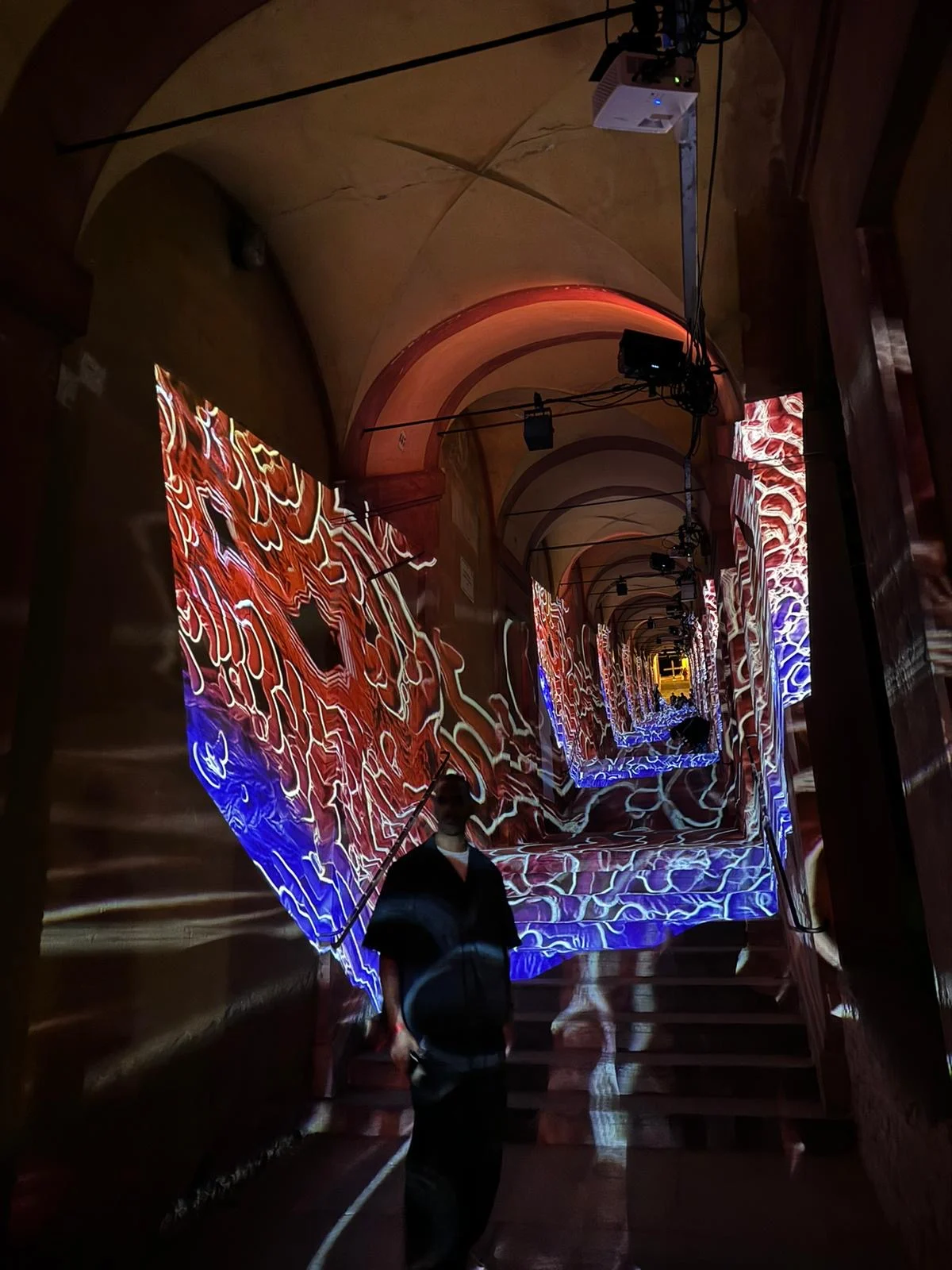 Proiezioni organiche rosse e blu che avvolgono le pareti del portico, proiettori appesi alla volta, un visitatore sotto – Cesare Cremonini Festival dei Portici, Bologna, Italia, 2024 – progettazione di eventi