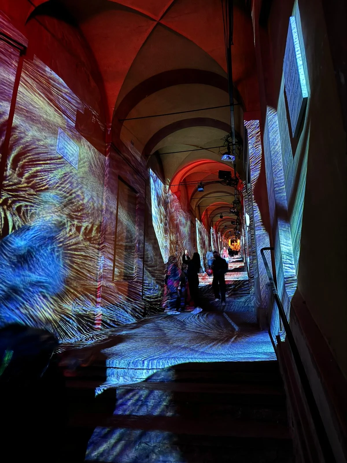 Portico di San Luca con proiezioni a vortici blu e oro su pareti e gradini, visitatori che lo attraversano – Cesare Cremonini Festival dei Portici, Bologna, Italia, 2024 – progettazione di eventi