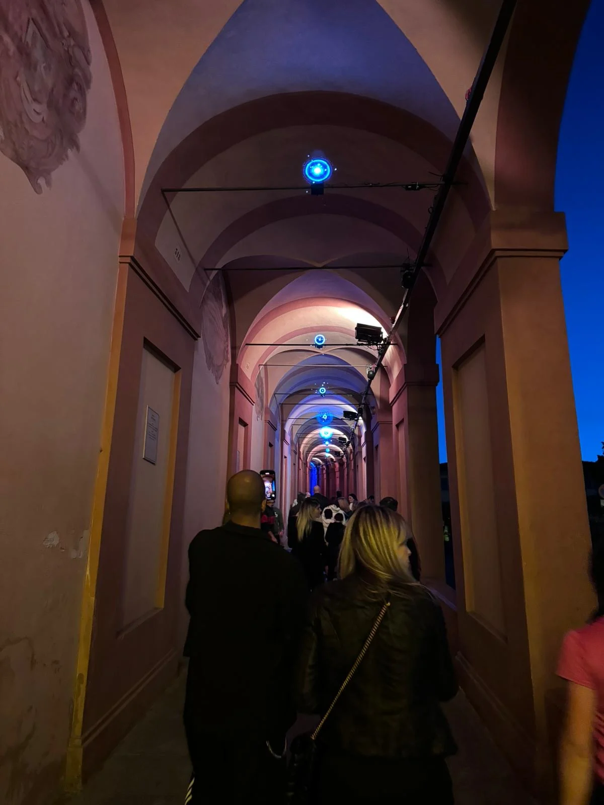 Visitatori che camminano sotto il portico al tramonto, volte illuminate di rosa con piccole luci blu lungo il soffitto – Cesare Cremonini Festival dei Portici, Bologna, Italia, 2024 – progettazione di eventi