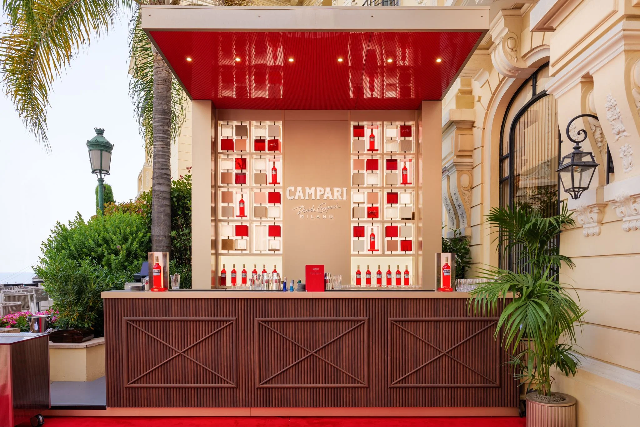Vista frontale del bar Campari: bancone in legno, scaffali illuminati di bottiglie rosse e una pensilina laccata rossa – Campari Pop-Up Bar all'Hôtel Hermitage, Monte Carlo, Principato di Monaco, 2024 – hospitality design