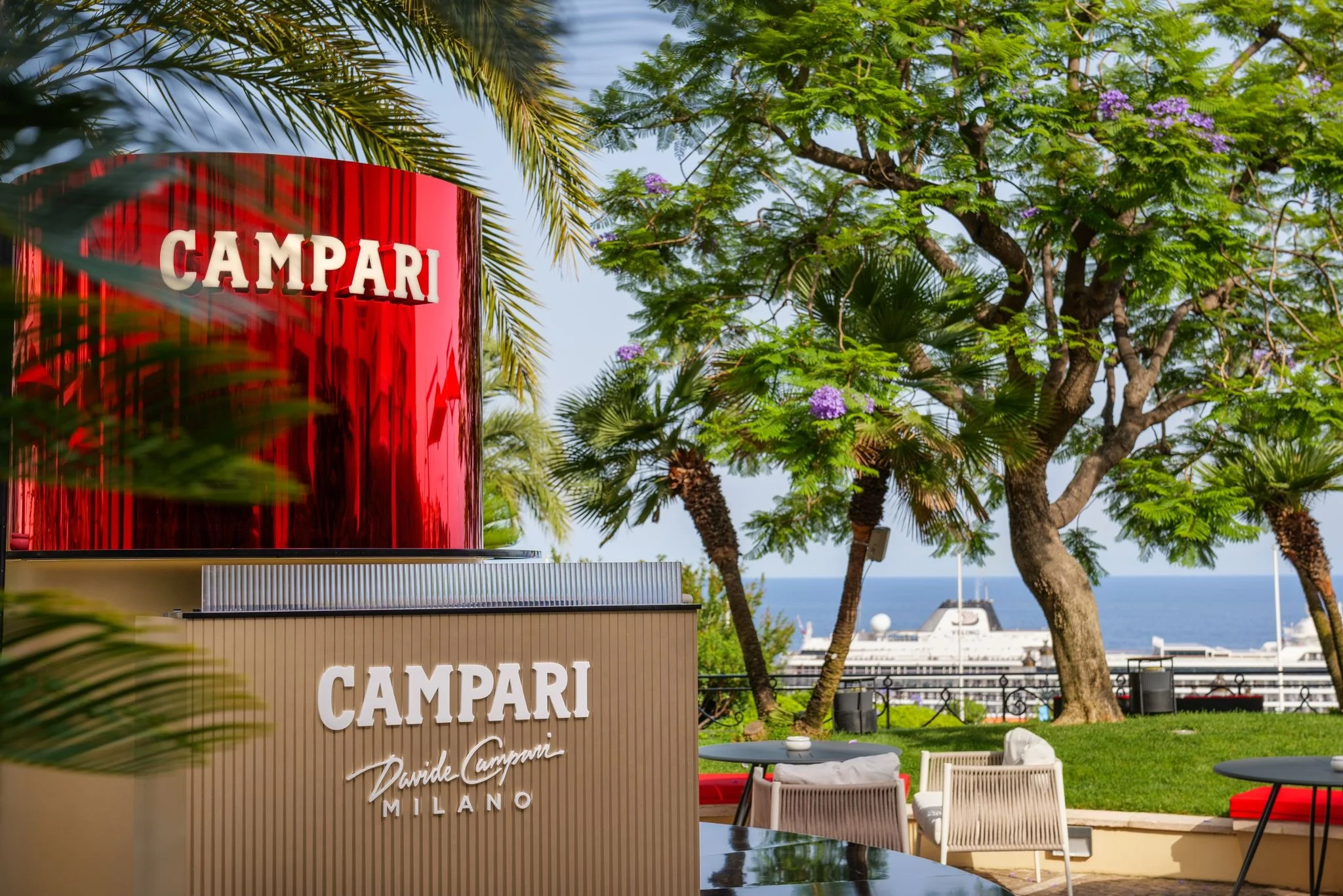 Tamburo a specchio rosso Campari sopra il bar, con sedute in giardino, alberi, il mare e il porto dietro – Campari Pop-Up Bar all'Hôtel Hermitage, Monte Carlo, Principato di Monaco, 2024 – hospitality design