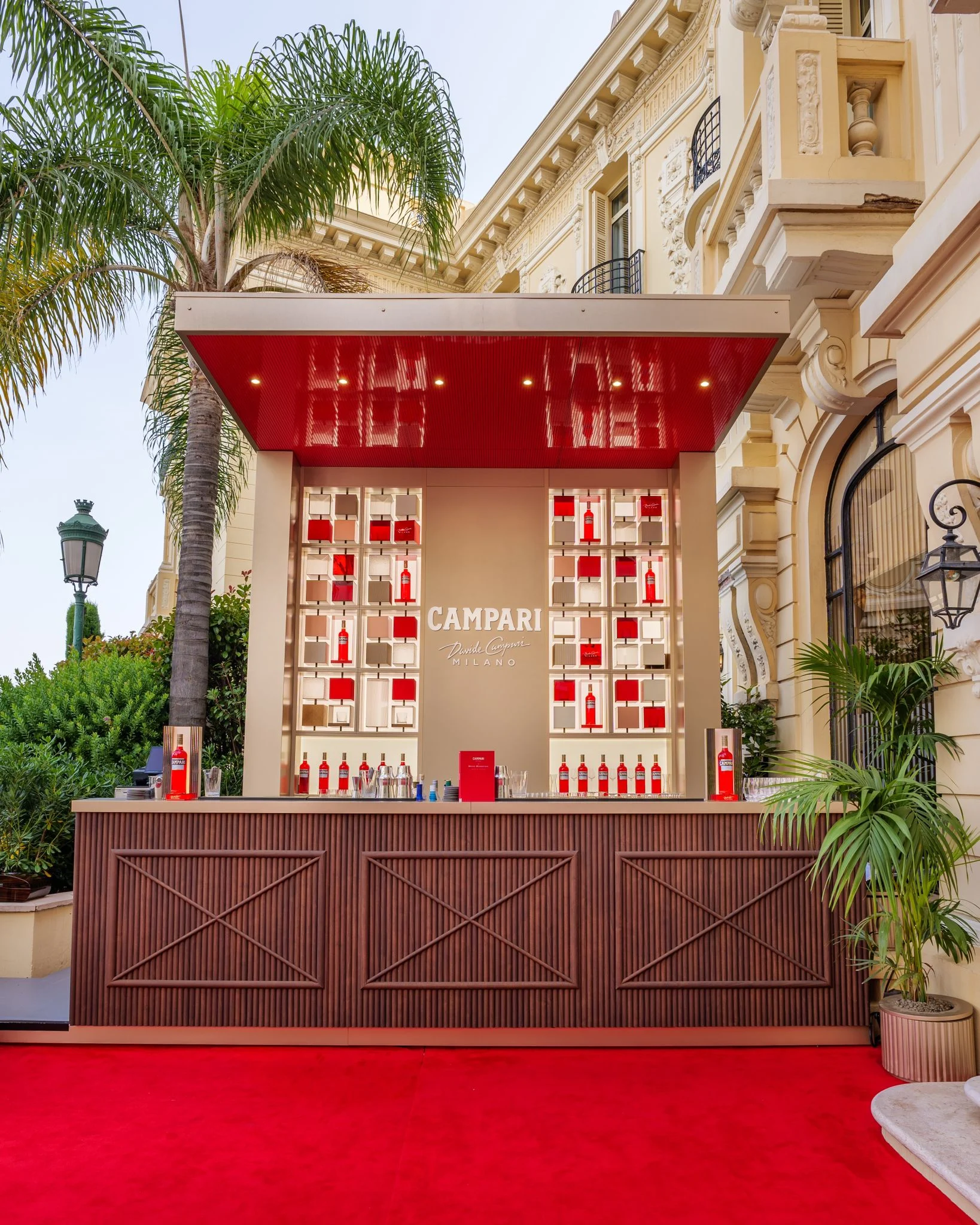 Pop-up bar Campari con pensilina rossa lucida, tappeto rosso e scaffali di bottiglie accanto alla facciata Belle Époque dell'hotel – Campari Pop-Up Bar all'Hôtel Hermitage, Monte Carlo, Principato di Monaco, 2024 – hospitality design