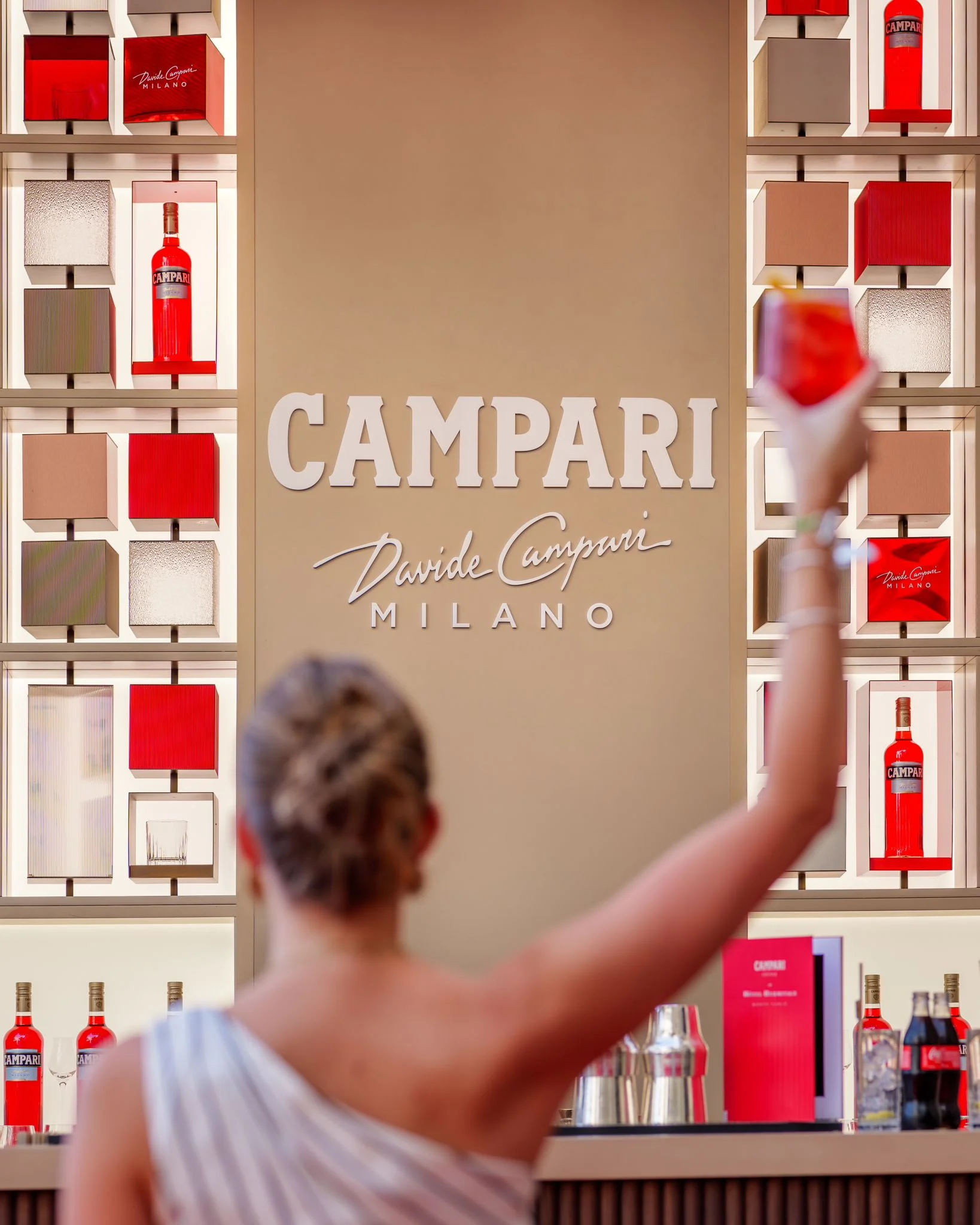 Ospite che alza un cocktail rosso davanti alla parete con il logo Campari Milano e scaffali di bottiglie – Campari Pop-Up Bar all'Hôtel Hermitage, Monte Carlo, Principato di Monaco, 2024 – hospitality design