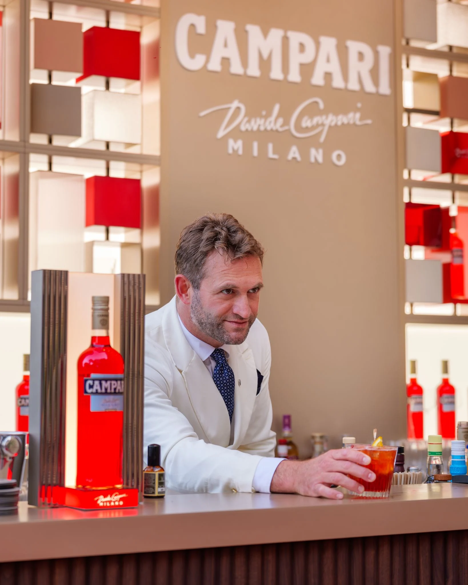 Barman in giacca bianca che serve un cocktail al bancone Campari, bottiglie rosse in esposizione – Campari Pop-Up Bar all'Hôtel Hermitage, Monte Carlo, Principato di Monaco, 2024 – hospitality design