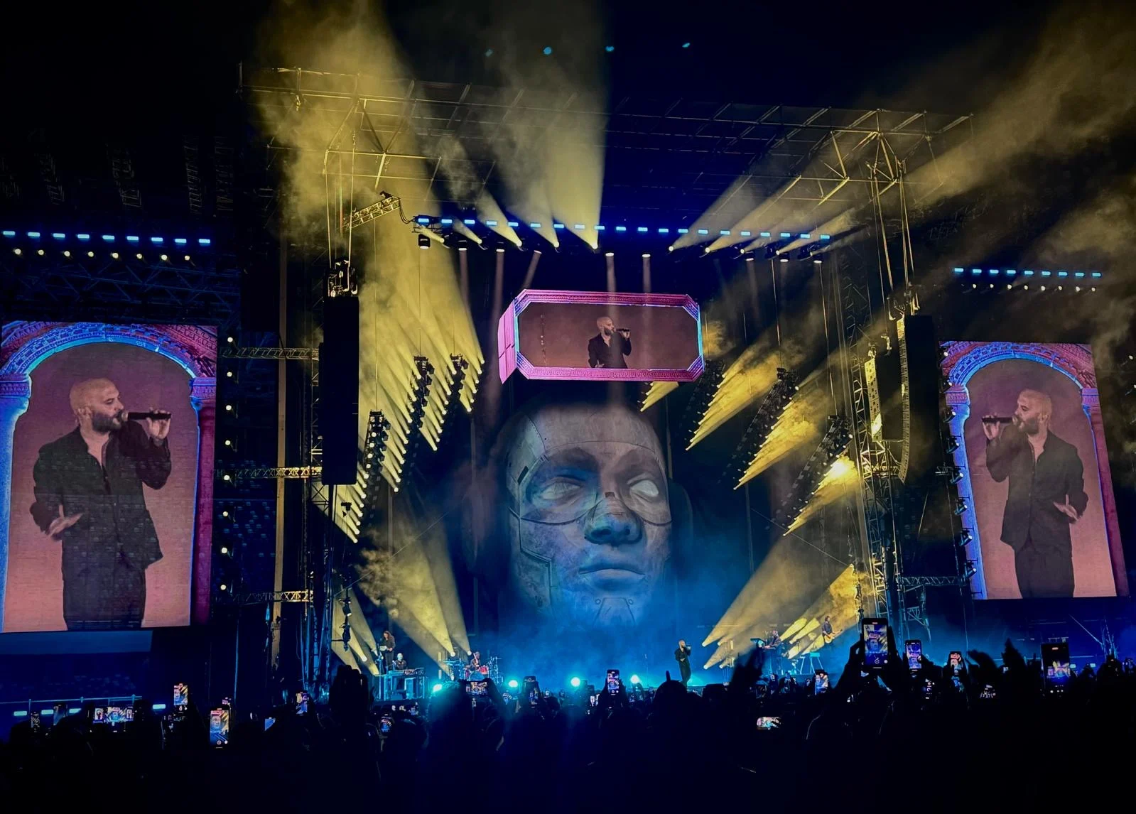 Palco negli stadi dei Negramaro: gigantesca testa scolpita con visiera-schermo, schermi LED laterali e fasci di luce gialli – Negramaro Stadi 2024, Italia – scenografia per concerti