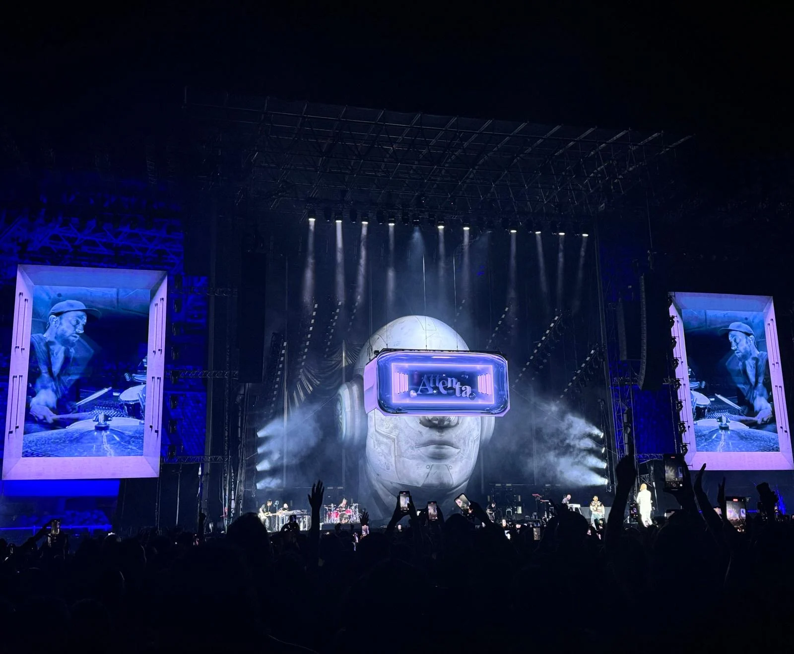 Vista d'insieme del palco nello stadio con la testa gigante con visiera, schermi laterali incorniciati e telefoni alzati – Negramaro Stadi 2024, Italia – scenografia per concerti