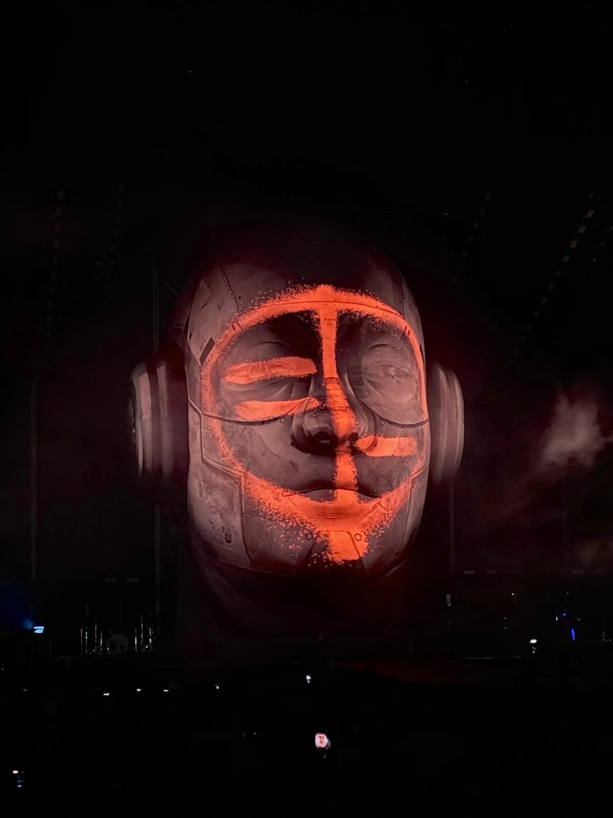 Scultura della testa gigante nel buio, con un video mapping arancione che traccia linee sul volto – Negramaro Stadi 2024, Italia – scenografia per concerti