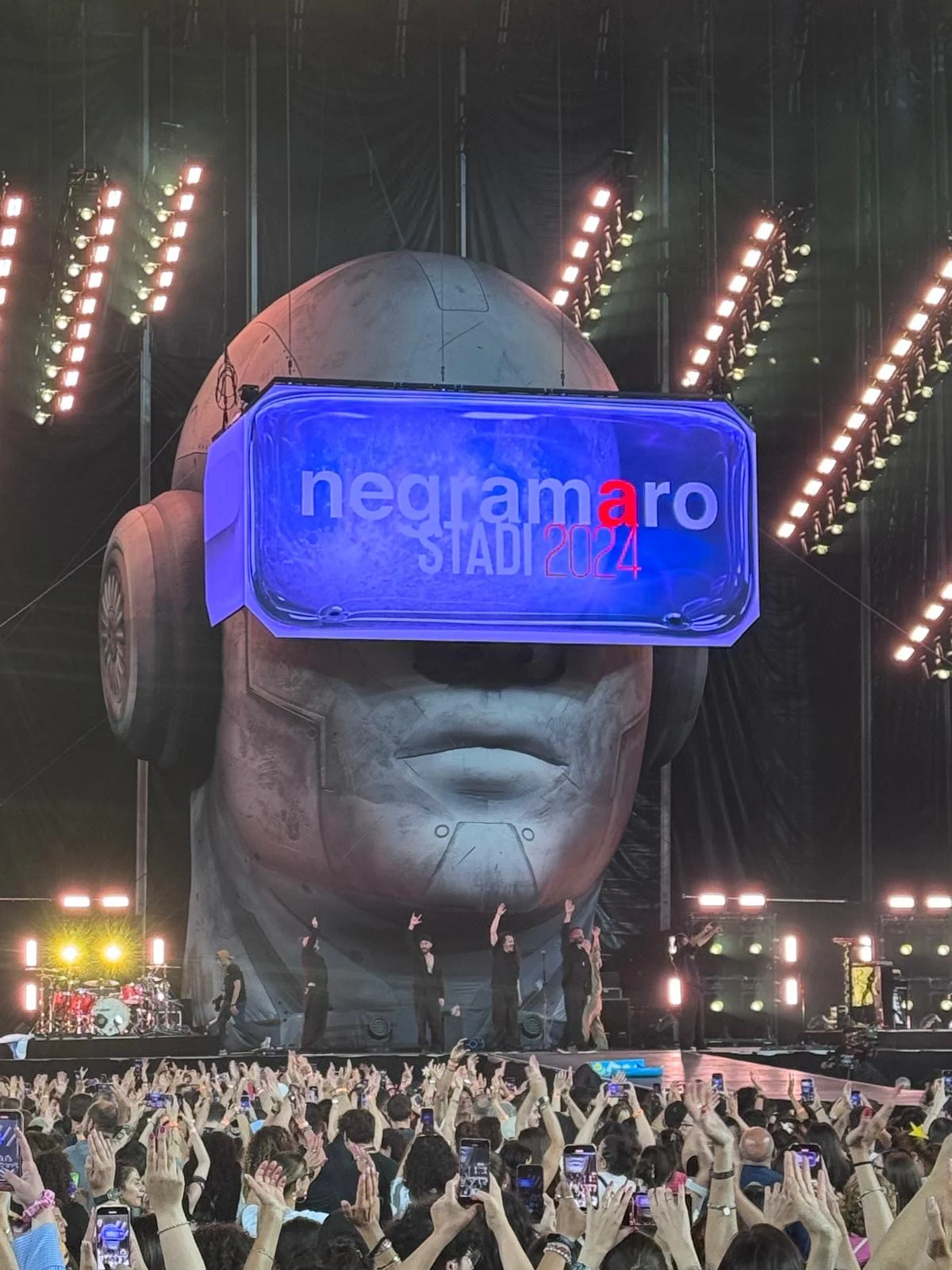 Testa gigante con cuffie e visiera LED con la scritta negramaro Stadi 2024, la band sul palco sotto – Negramaro Stadi 2024, Italia – scenografia per concerti