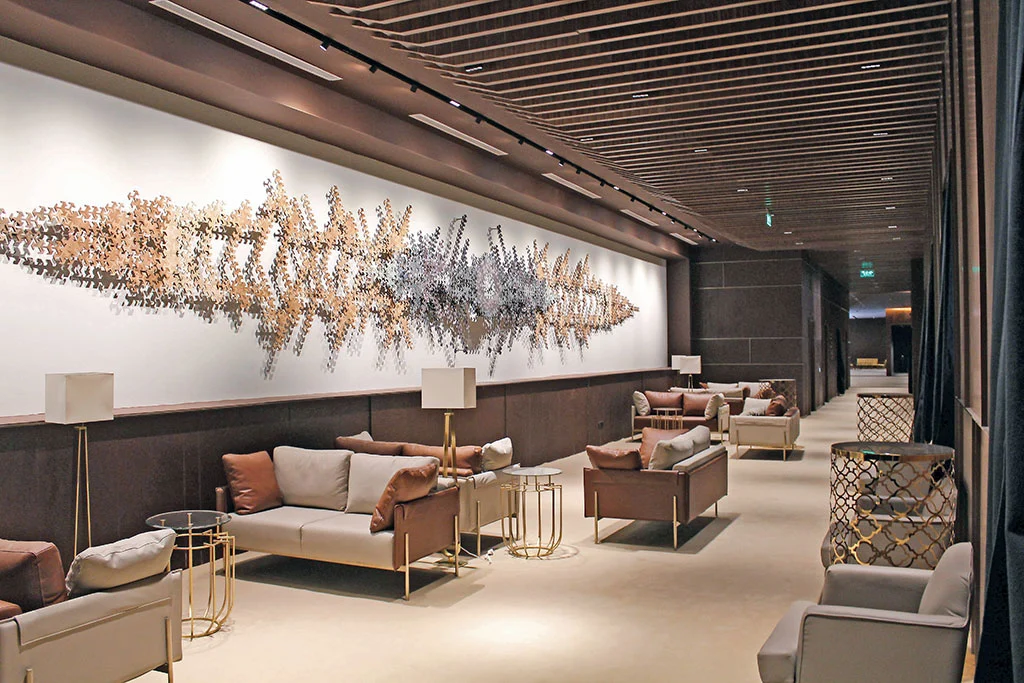 Lounge con divani in pelle, tavolini dorati e una lunga scultura metallica a parete sotto un soffitto a doghe – Maraya Concert Hall, AlUla, Arabia Saudita, 2019 – architettura